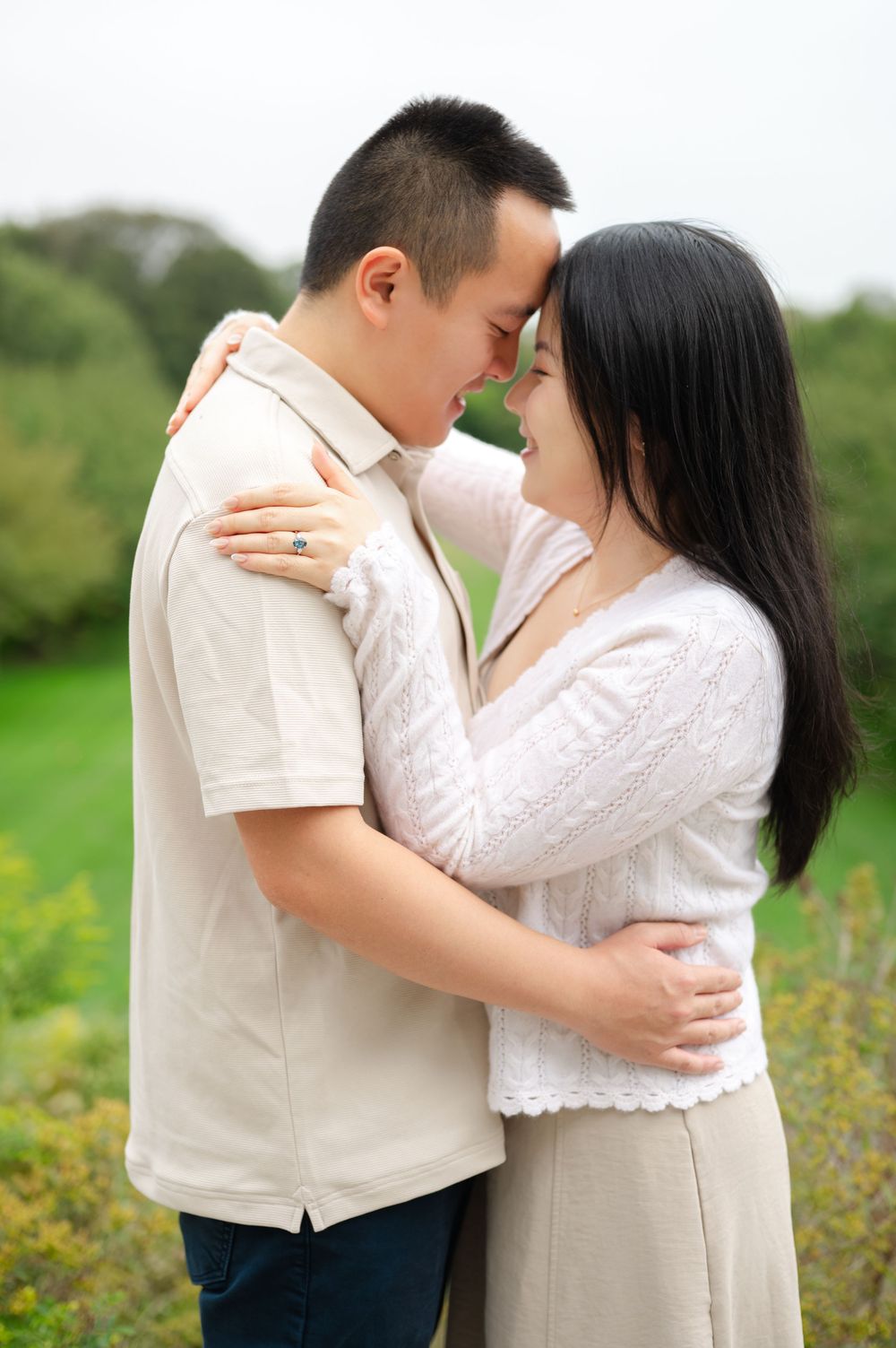 Asian couple embracing forehead-to-forehead, smiling lovingly in an outdoor garden setting.