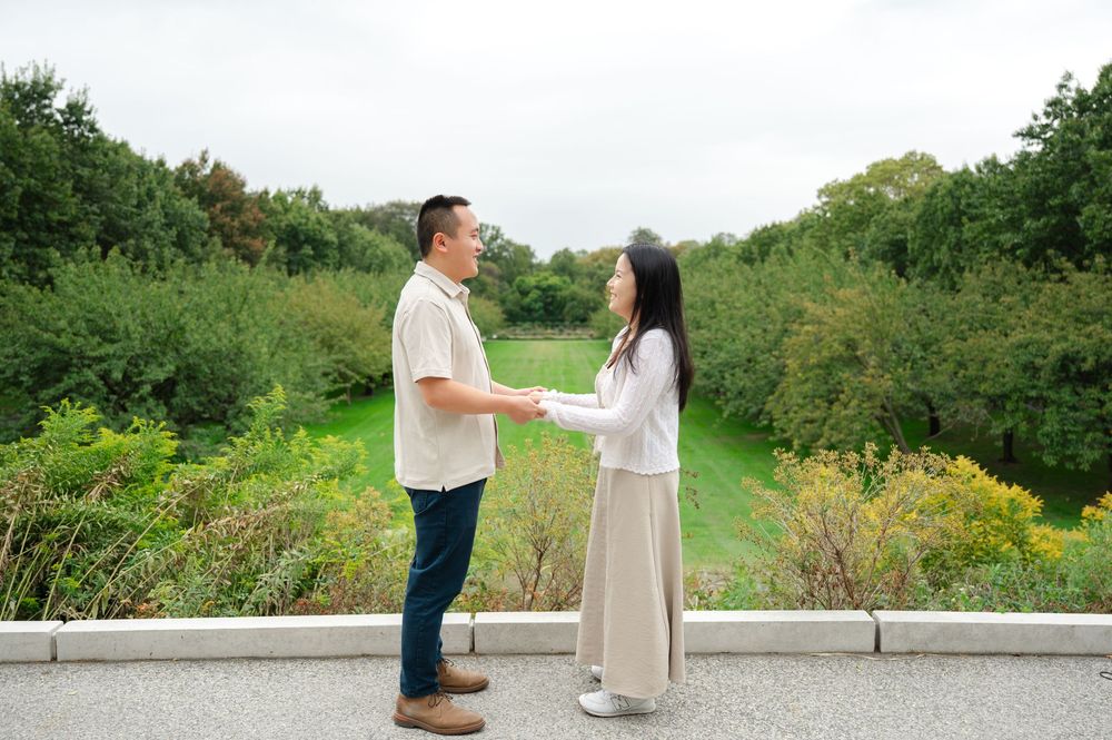 A couple holds hands facing each other on a path surrounded by green trees in a park.