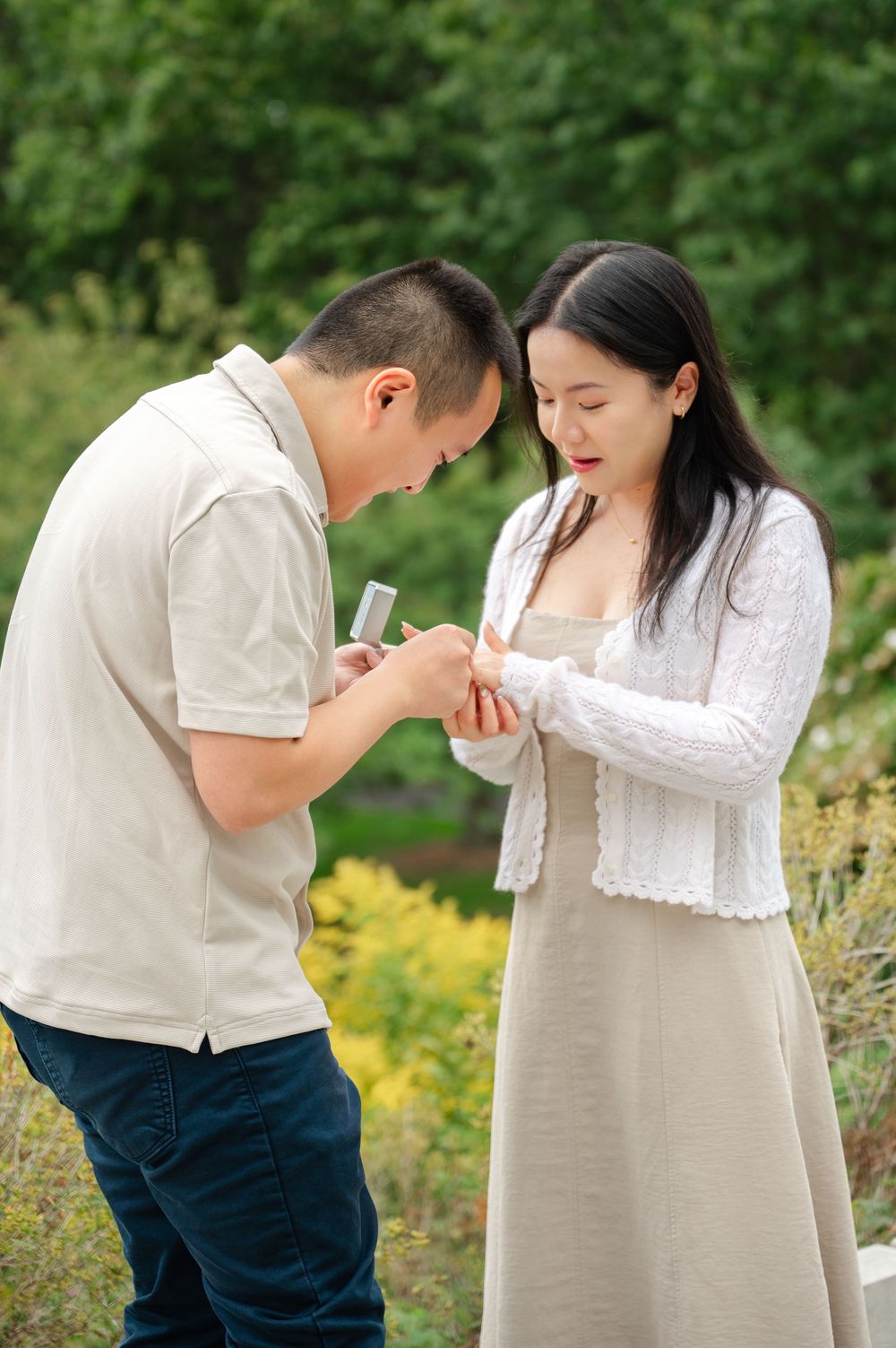 Man placing an engagement ring on his surprised girlfriends finger during an outdoor proposal in a garden.