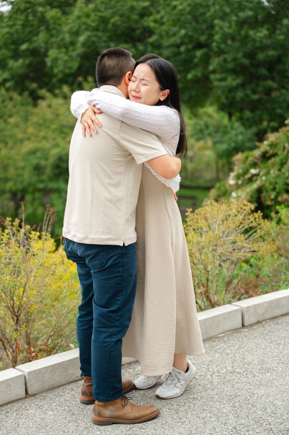 A loving Asian couple shares a warm embrace outdoors in a lush green park setting.