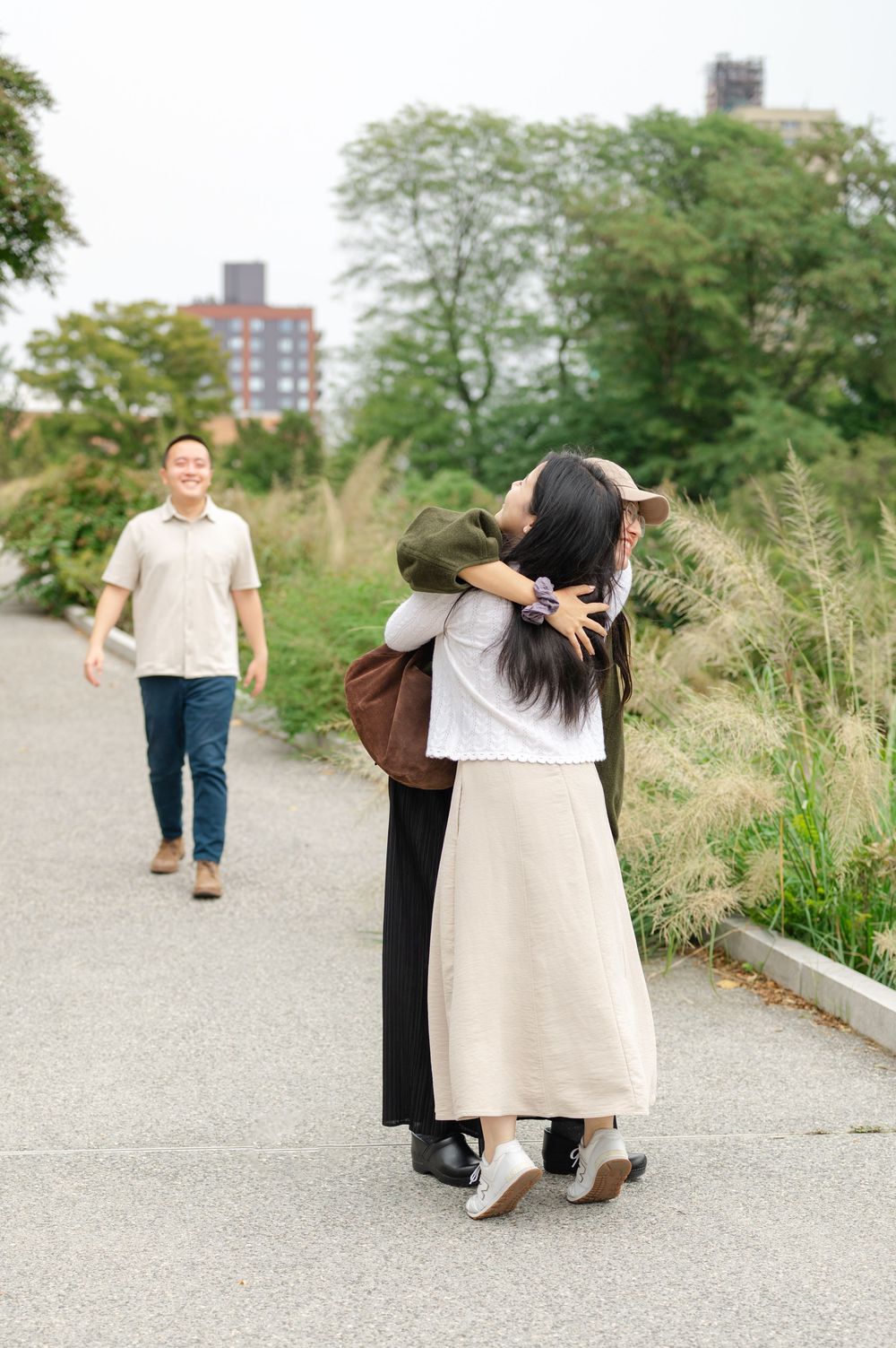 Asian woman hugging a friend on a garden path while her partner walks toward them.