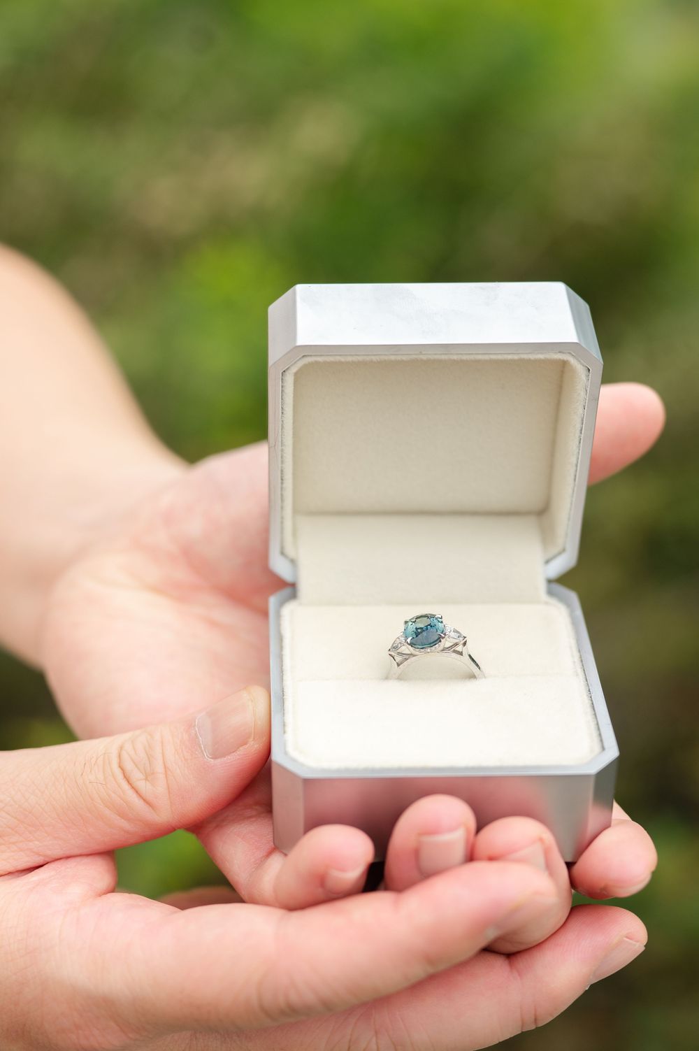 Hands holding an open gray jewelry box with a silver ring featuring a blue gemstone, set against a green outdoor background.