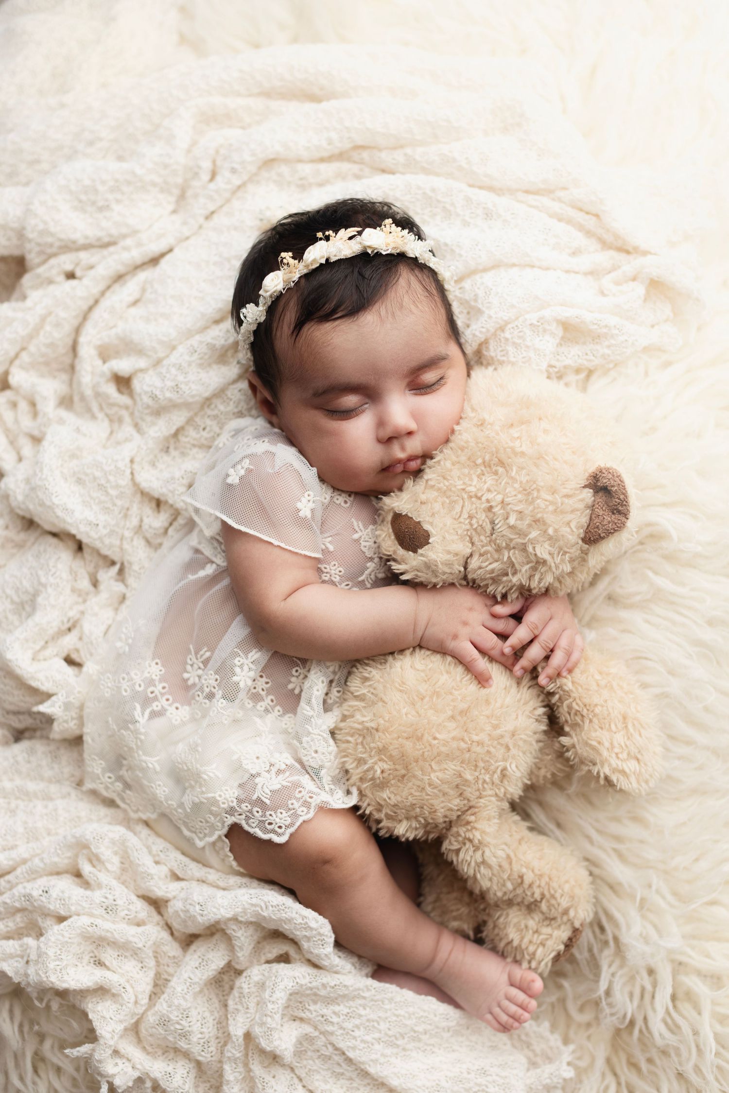 A woman in a floral dress holds a sleeping baby in a white lace outfit against a soft studio background.