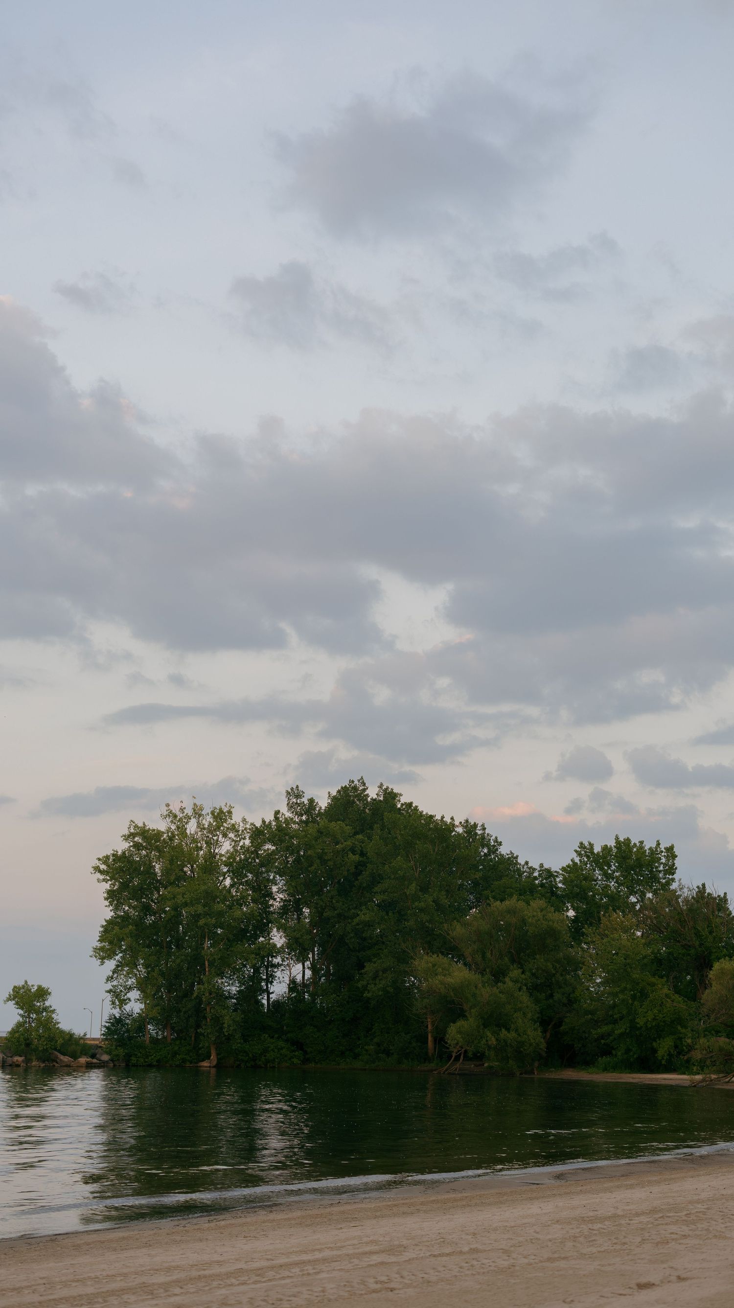 Serene lakeside view with lush green trees on a small island, sandy shore, and dramatic cloudy sky at dusk.