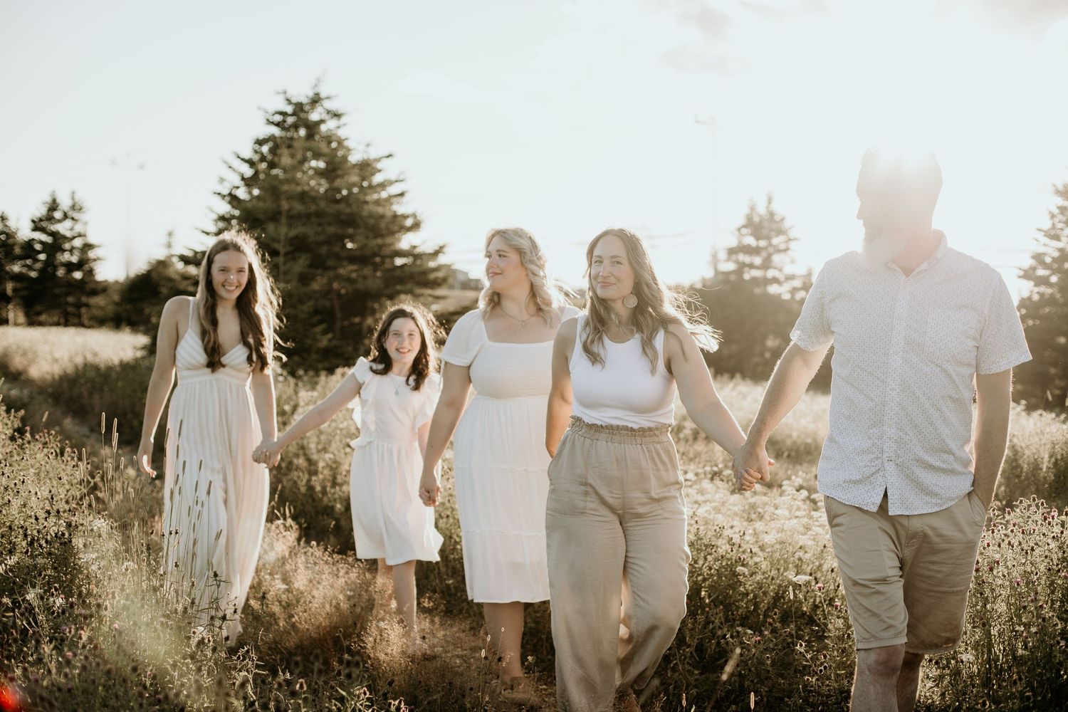 family of 5 with 3 girls holding hands walking through a field with sun shining through the trees
