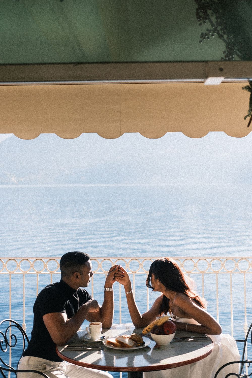 People sit at a table overlooking a scenic lake view through white railings on a sunny day.