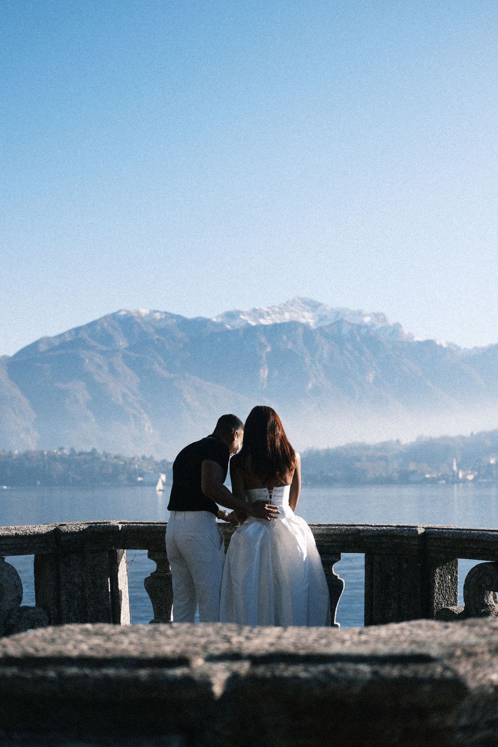 A silhouetted couple sits on a stone wall overlooking a scenic mountain landscape with snow-capped peaks against a blue sky.