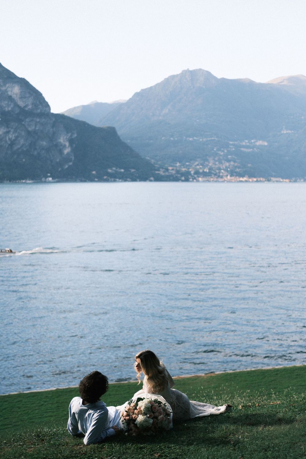 Couple sitting on green grass by a serene lake with mountain backdrop, bride holding bouquet in white dress.