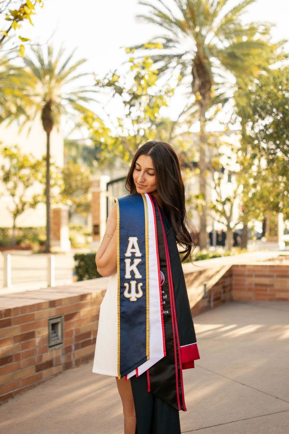 Graduate poses with Alpha Kappa Psi stole and honor cords outdoors near palm trees.