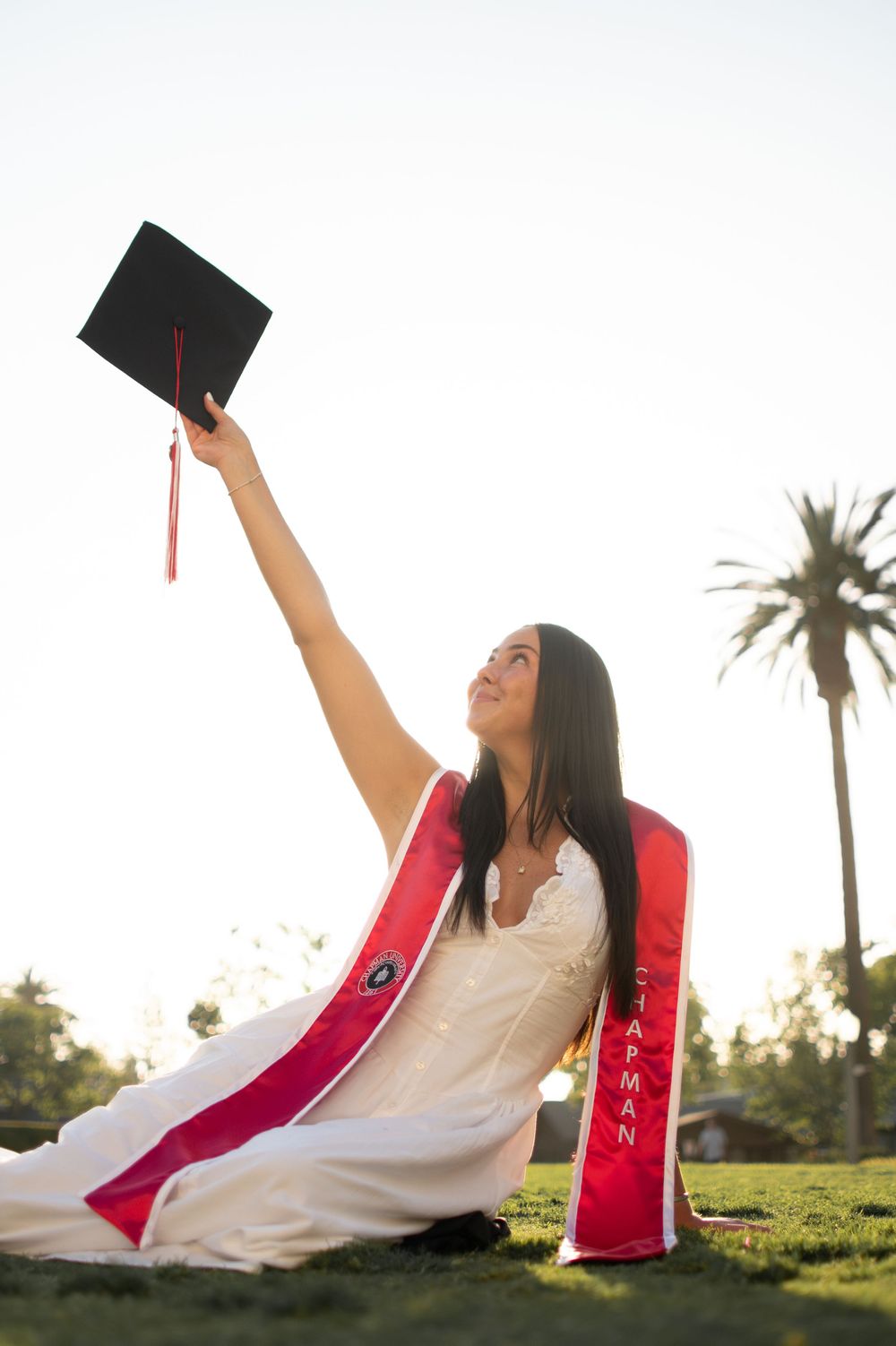 Graduate raises mortarboard cap triumphantly while seated on grass wearing Chapman University sash.