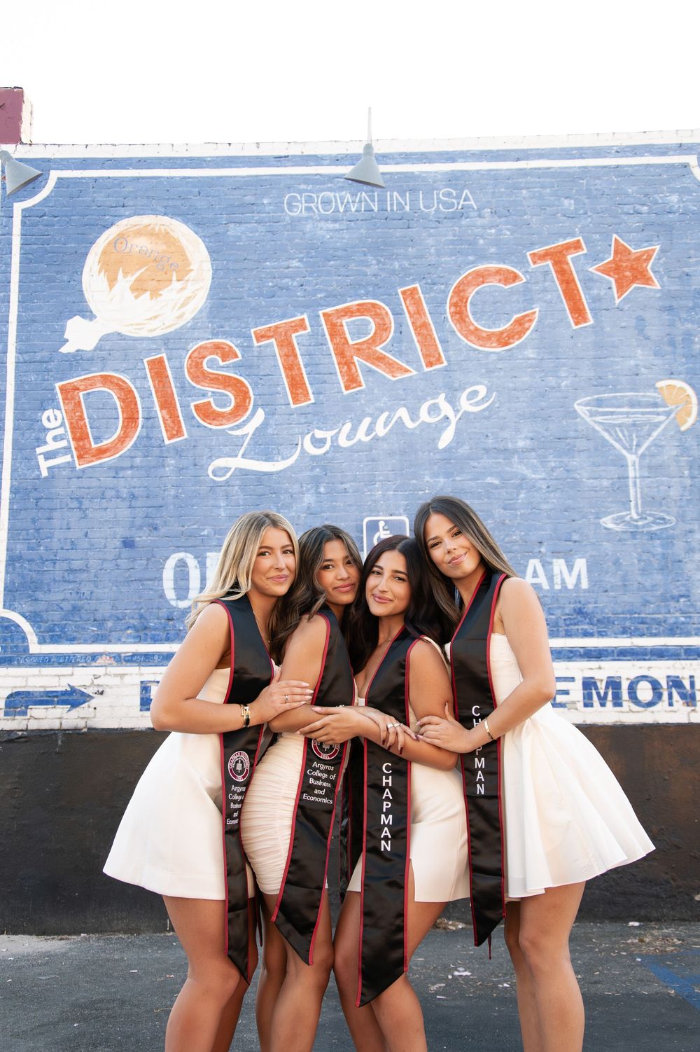 Four women in white dresses with Chapman graduation sashes pose at The District Lounge.