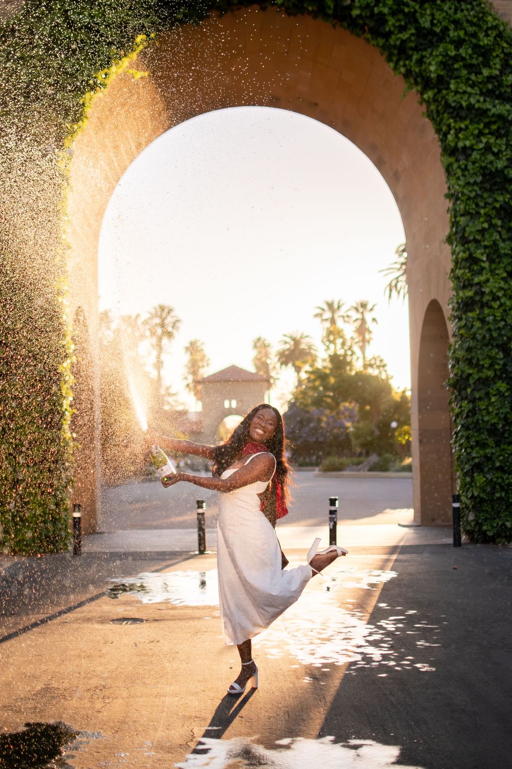 Joyful woman in white dress pops champagne under ivy-covered arch at golden hour.