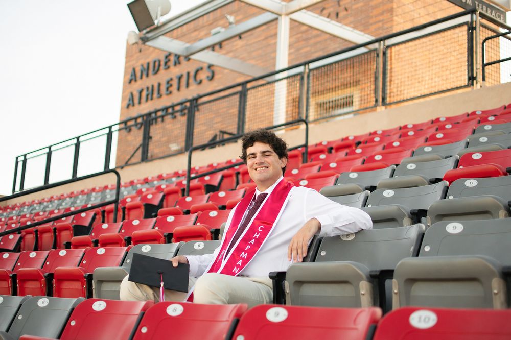 Male graduate sits in stadium bleachers wearing red sash near Ander Athletics Center sign.