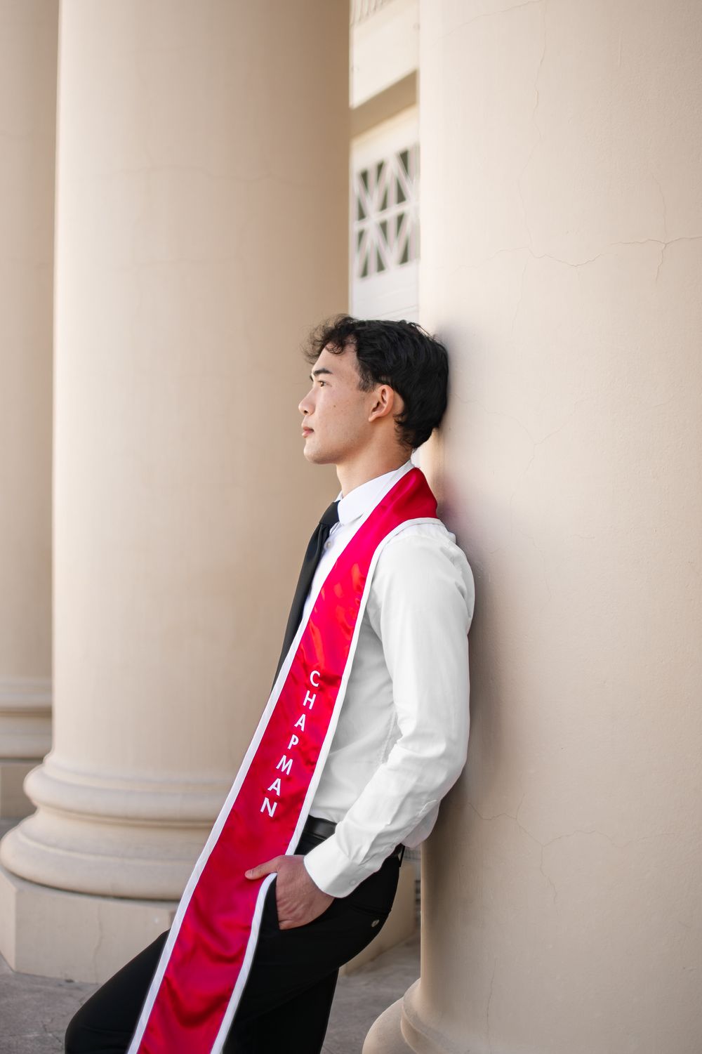 Young male Chapman University graduate leans against white column wearing red graduation sash and formal attire.