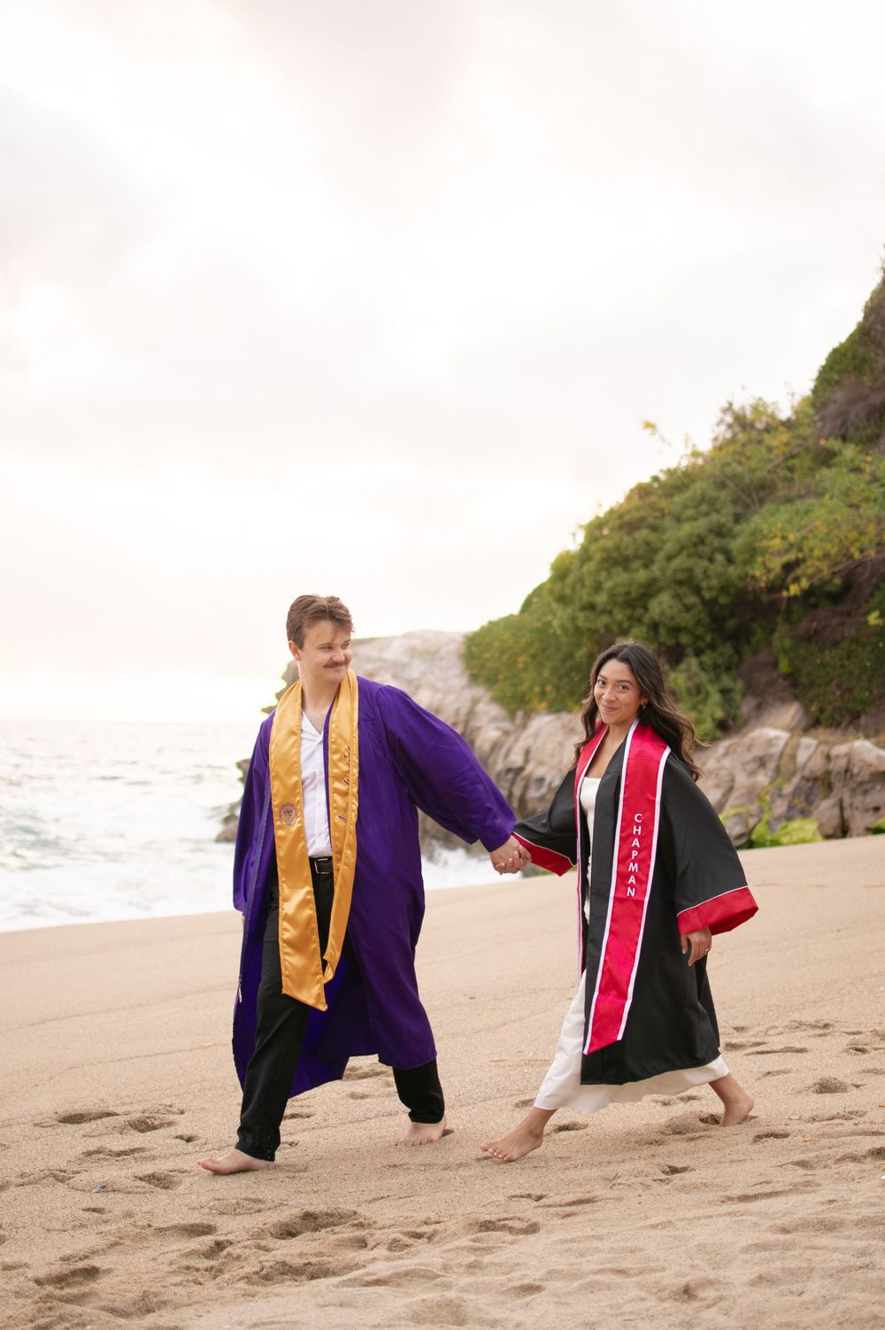 Two graduates in robes holding hands walking barefoot on a scenic beach.