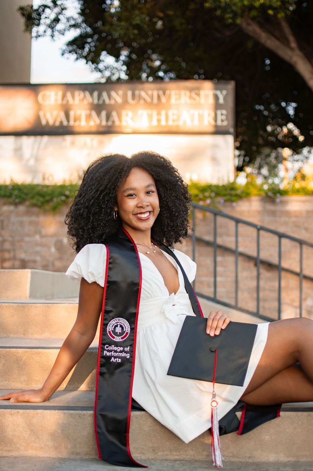 Smiling graduate in white dress holds cap and College of Performing Arts sash at Chapman University Waltmar Theatre.