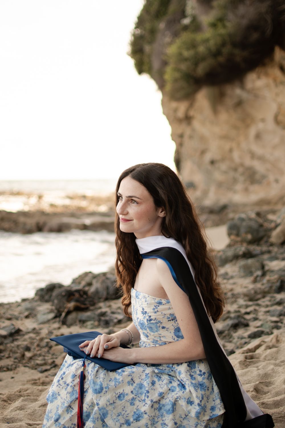 Contemplative female graduate in floral dress holds diploma cover while sitting on scenic rocky beach at sunset.