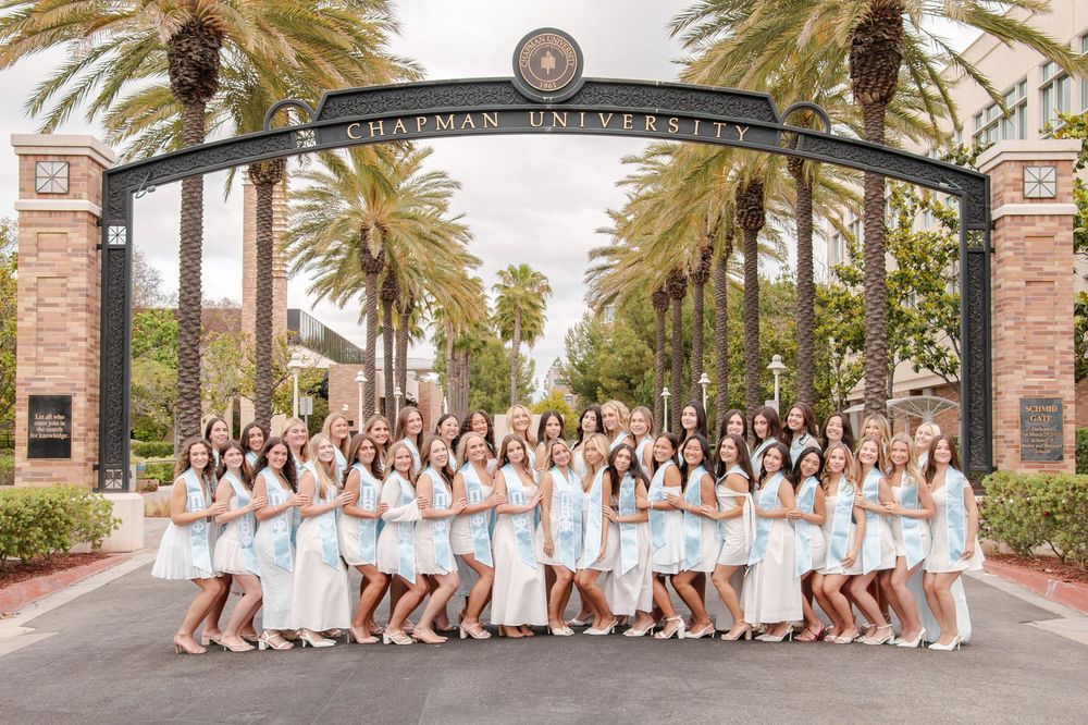 Large group of women in light blue and white dresses pose under Chapman University arch.