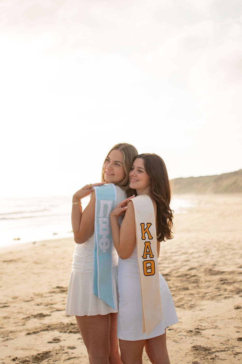 Two college women in white dresses wear sorority sashes KAO and DPhi on a sunny beach.