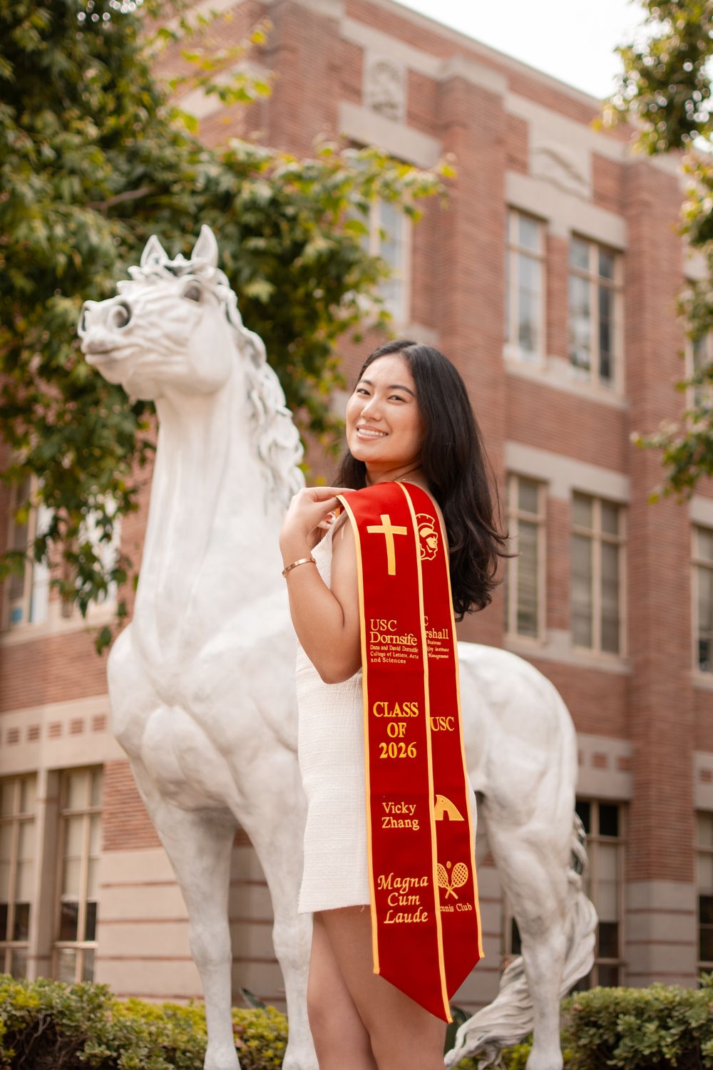 USC graduate Vicky Zhang poses with white horse statue wearing Class of 2016 Magna Cum Laude red graduation sash.