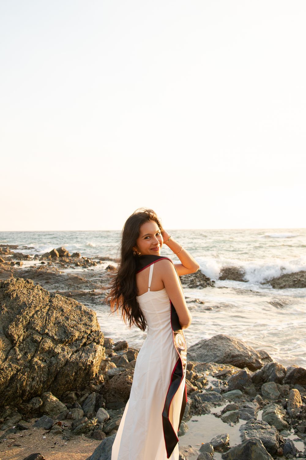 Young woman in white dress stands on rocky beach at sunset holding a colorful sash.