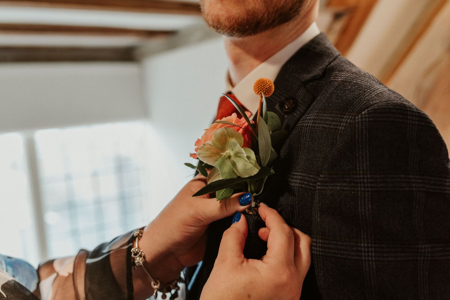 Someone adjusts a pink and green floral boutonniere on a dark plaid suit jacket at a formal event.