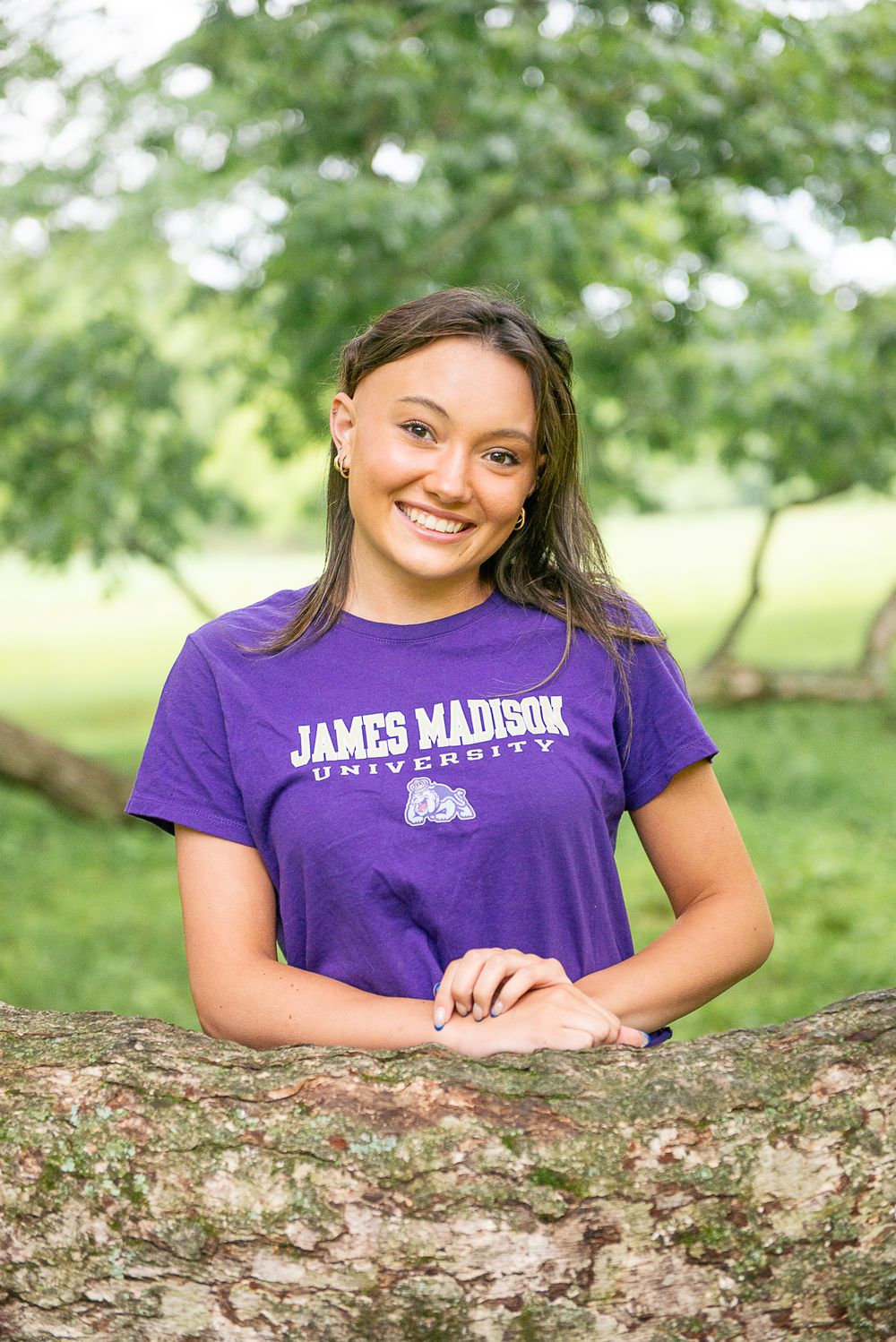 Cheerful outdoor portrait in purple James Madison University shirt leaning on tree trunk in park setting.