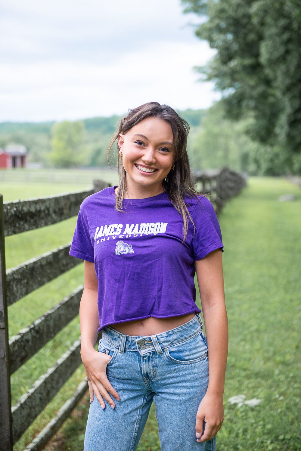 Casual outdoor portrait in purple crop top and jeans leaning against rustic wooden fence.