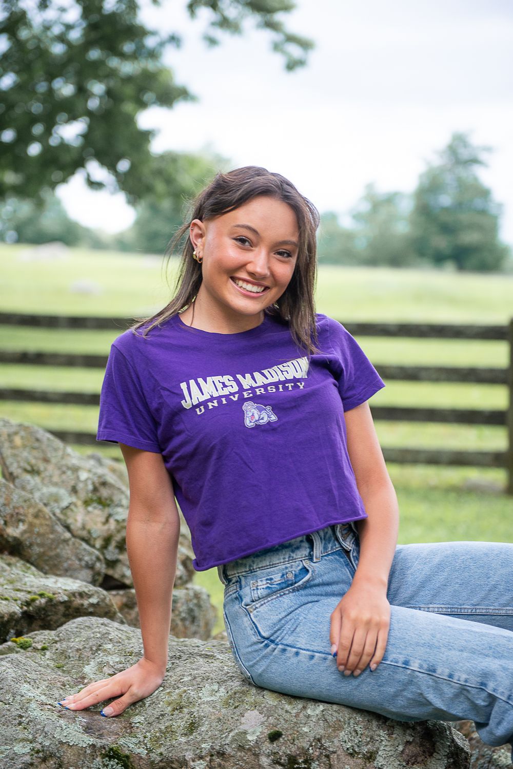 Relaxed portrait on rock wearing purple James Madison University shirt and jeans with fence in background.
