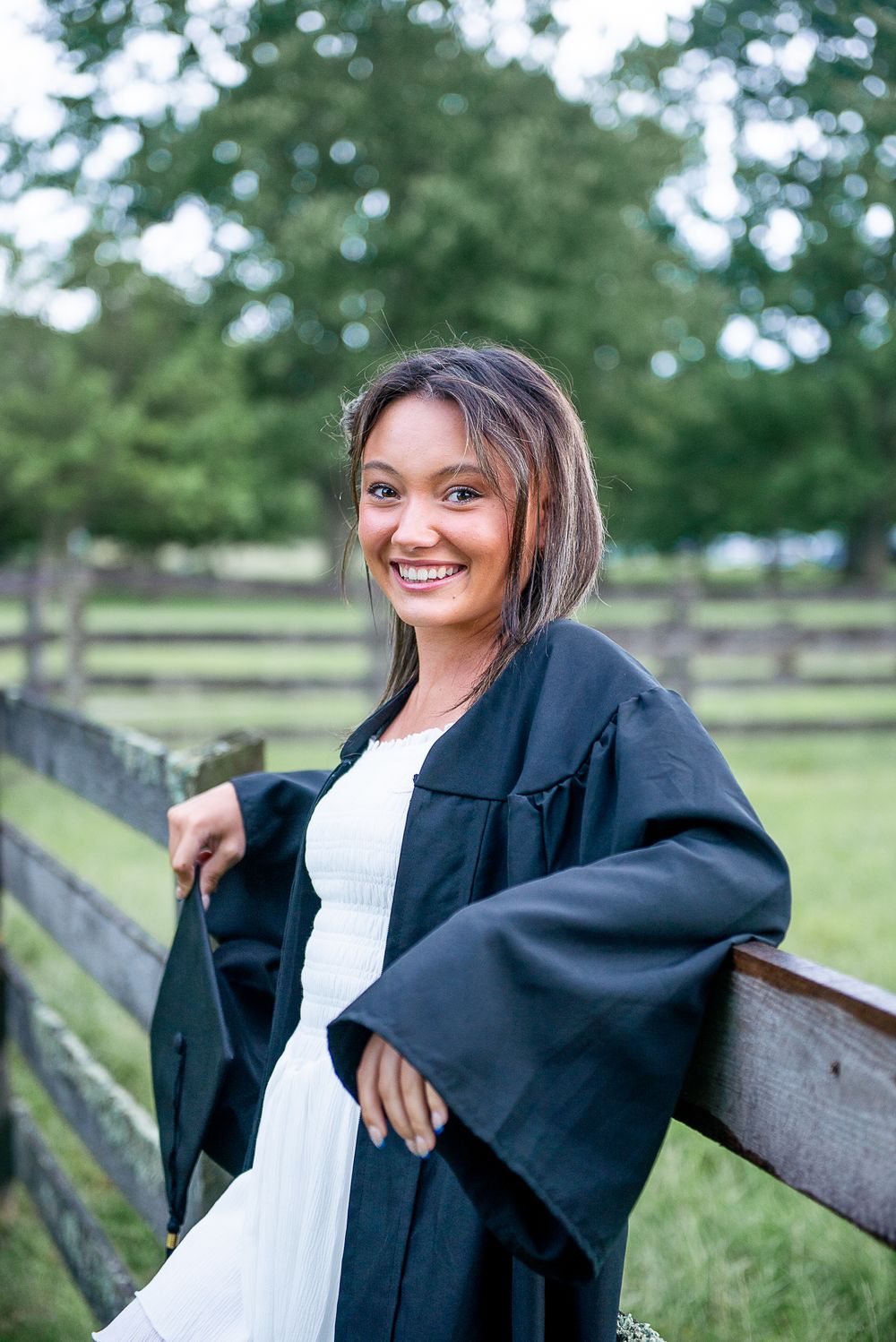 Graduate portrait in black gown and white dress leaning on wooden fence with pastoral background.