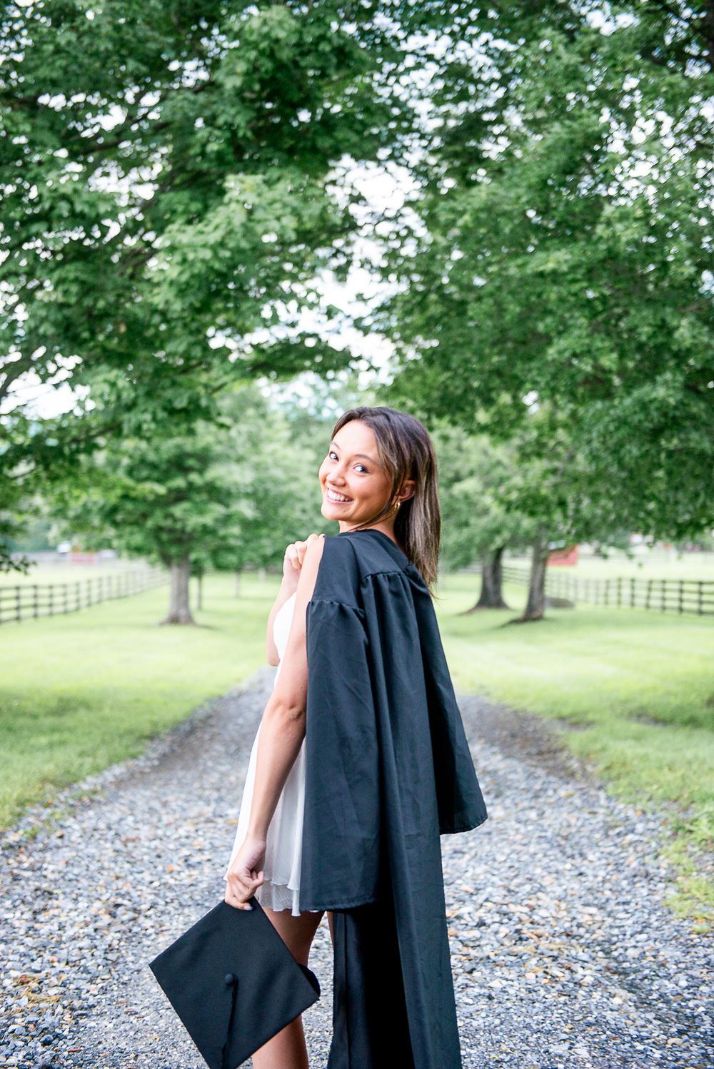 Graduate poses on a gravel path wearing academic regalia and white dress against a backdrop of green trees.