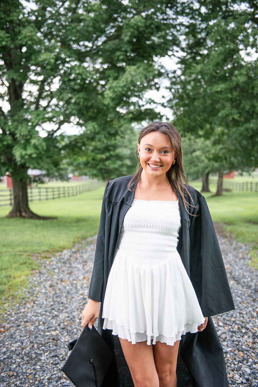 Graduation photo on gravel path wearing white dress and black gown with trees in background.