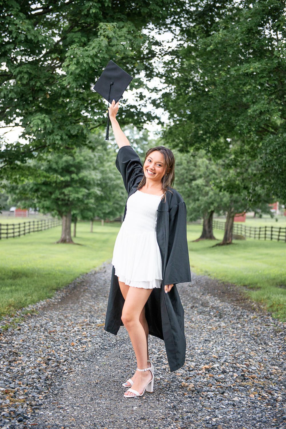 Joyful graduate in white dress raises cap high on rustic path surrounded by green trees.