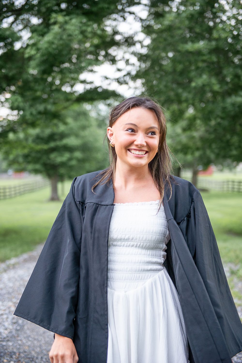 Graduate smiles brightly wearing academic regalia and white dress on scenic campus path.