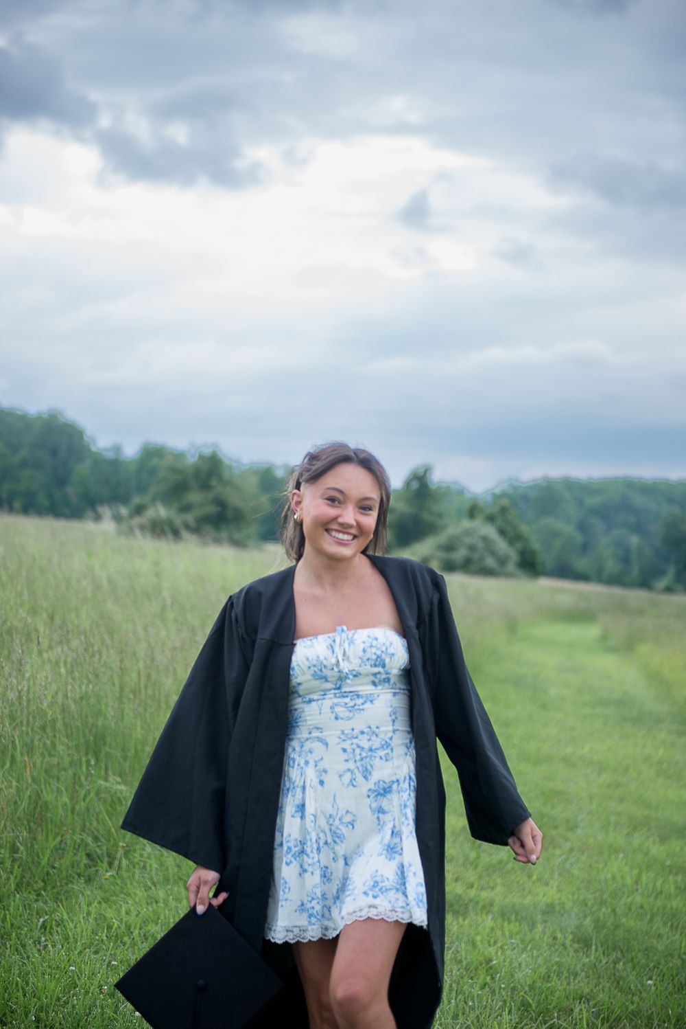 Graduate walks through meadow wearing black academic gown and blue floral dress.