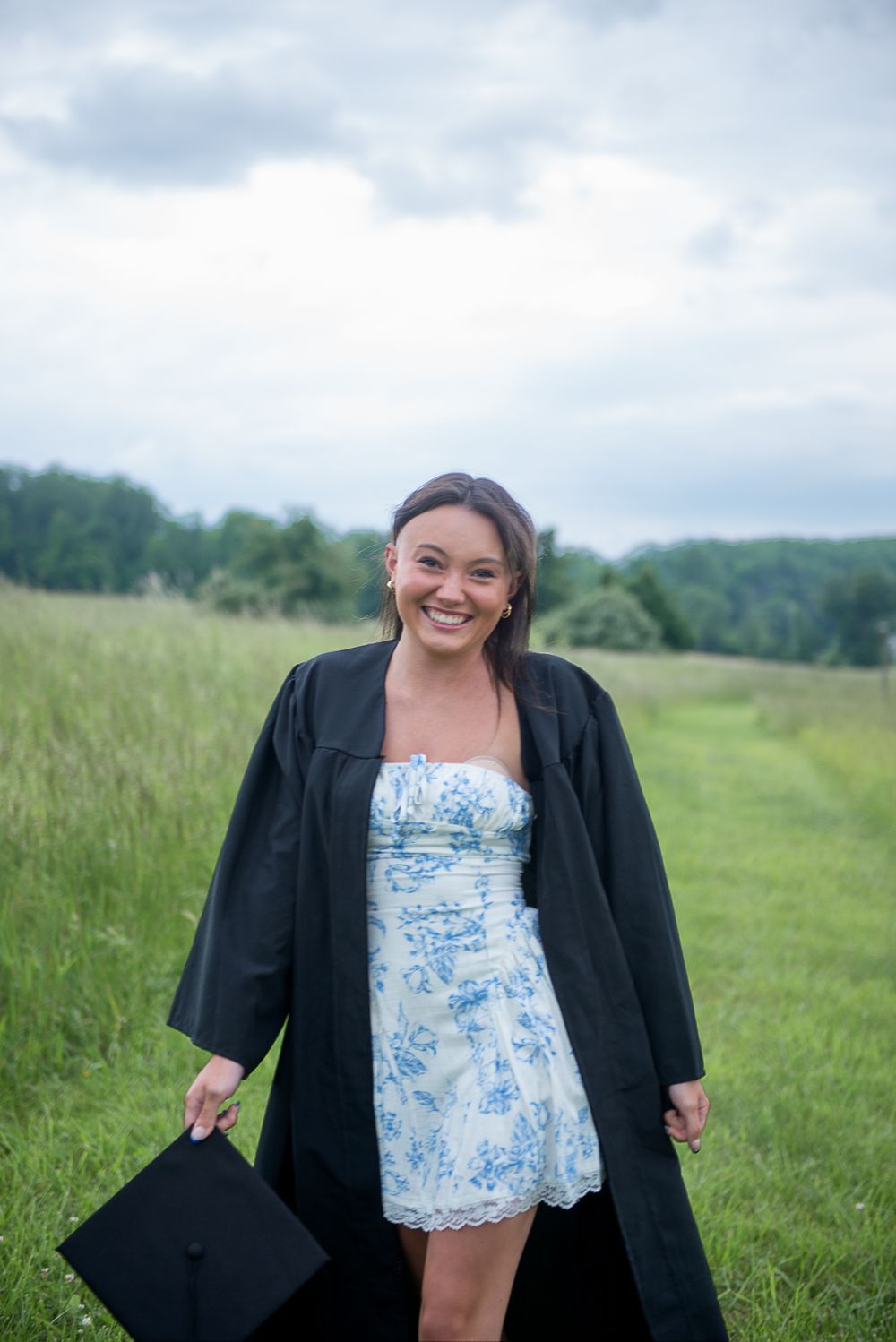 Graduate holds cap while wearing floral dress and academic gown in pastoral field setting.