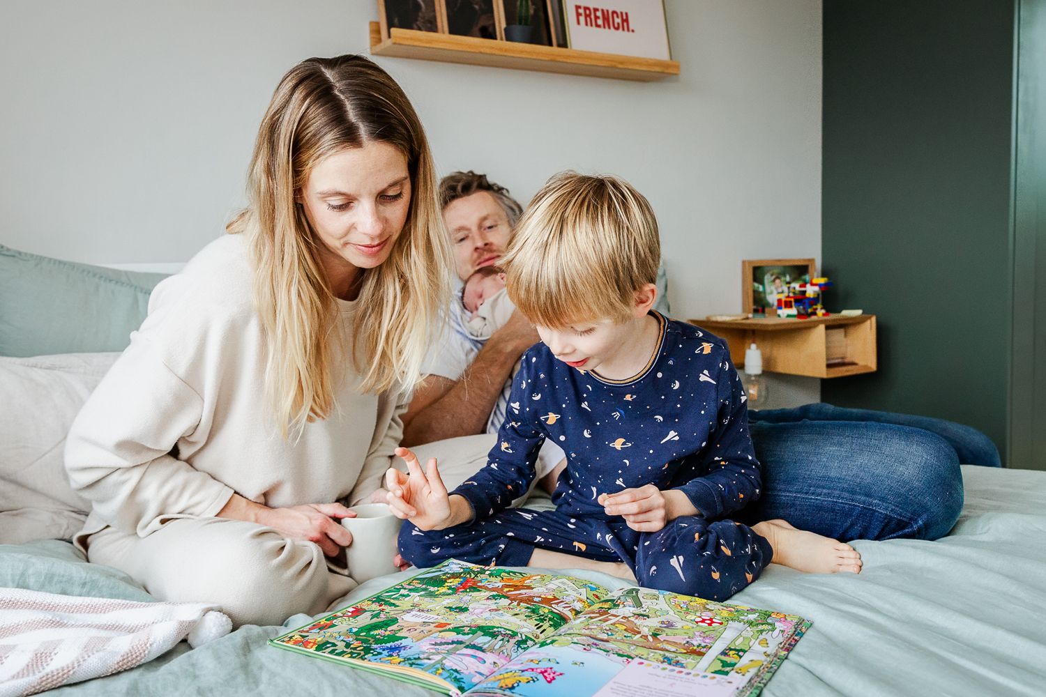 A family read a book on the bed at their newborn photoshoot. Dad looks relaxed as he holds the baby. Bristol natural motherhood photographer