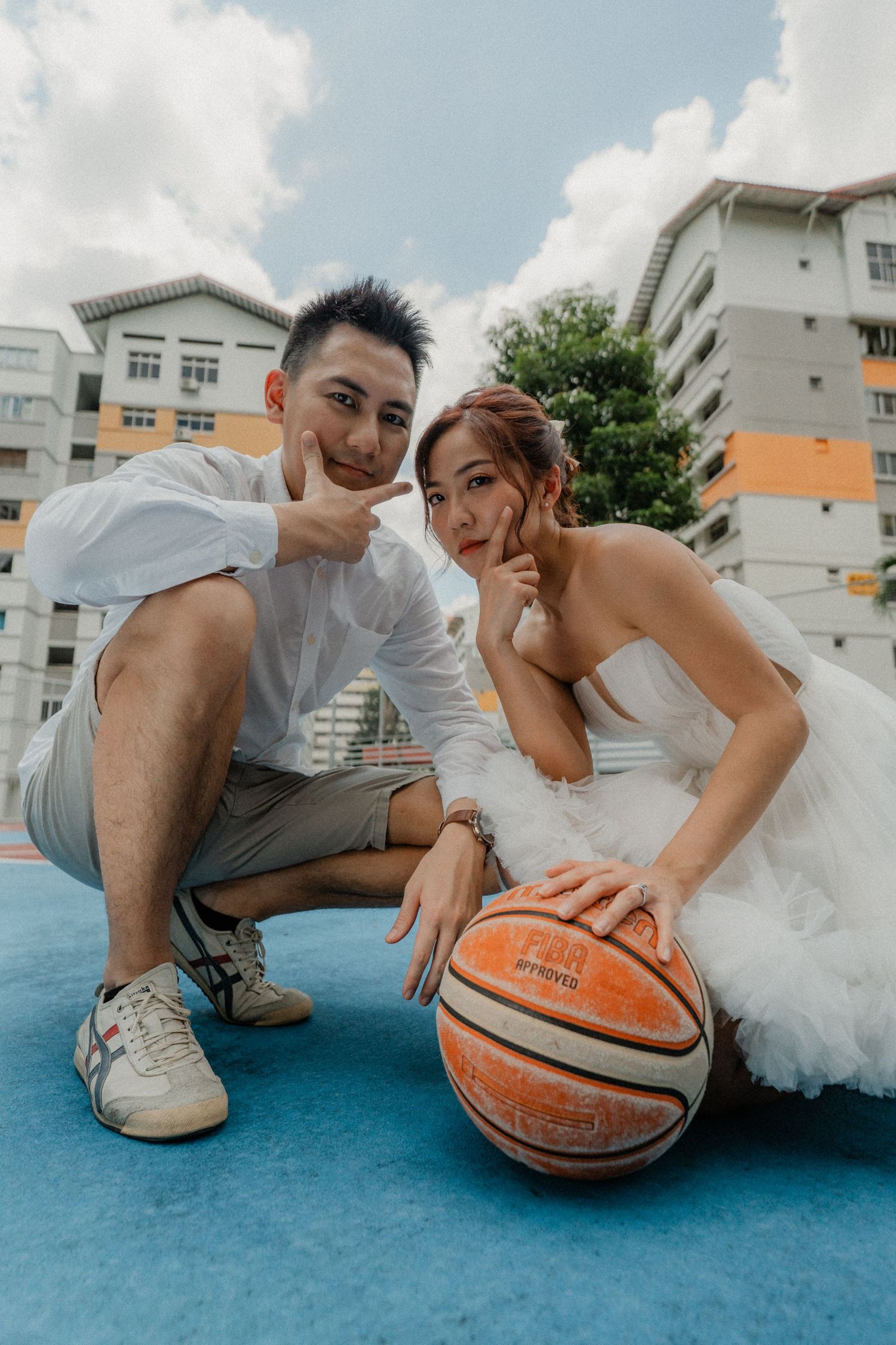 A couple in wedding attire pose together with a basketball on a blue court outdoors near apartment buildings.