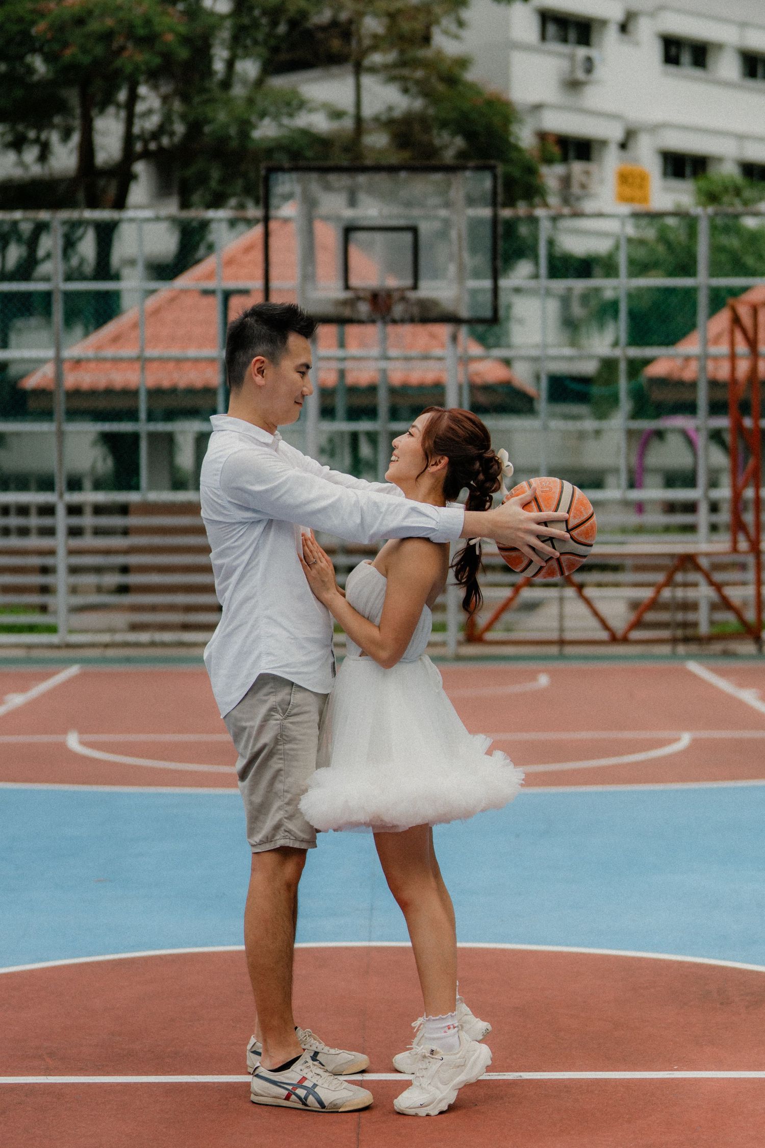 A couple playfully poses with a basketball on an outdoor court, with the person in a white dress bending backwards.