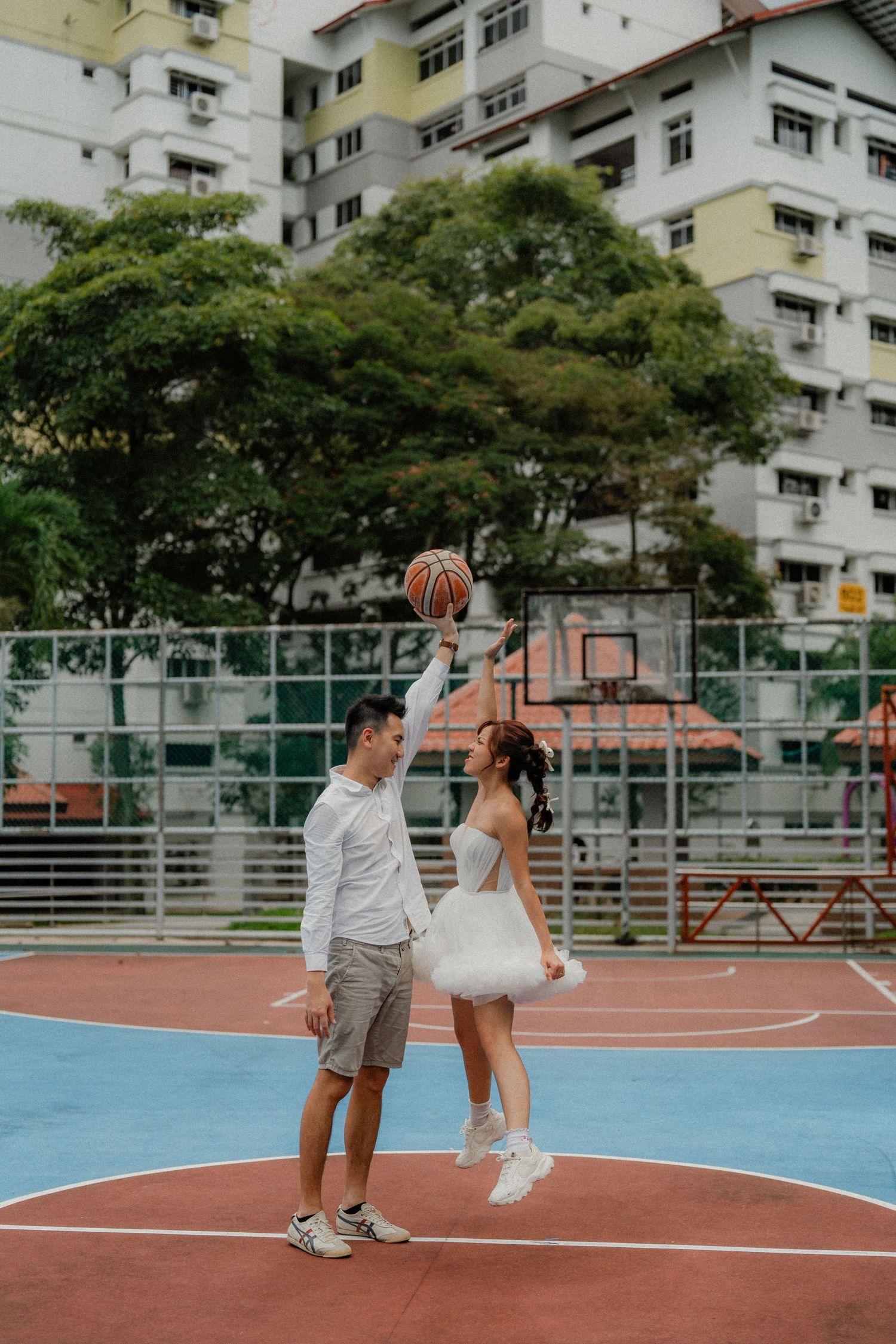 A couple shares a playful basketball moment on an urban court surrounded by residential buildings.