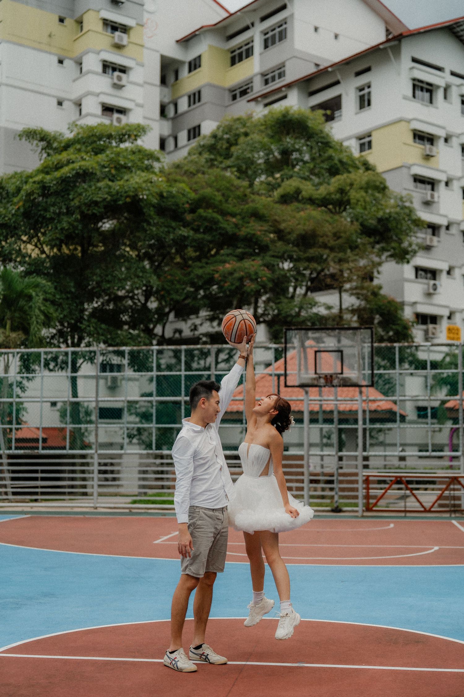 A couple plays basketball together on an outdoor court with apartment buildings in the background.