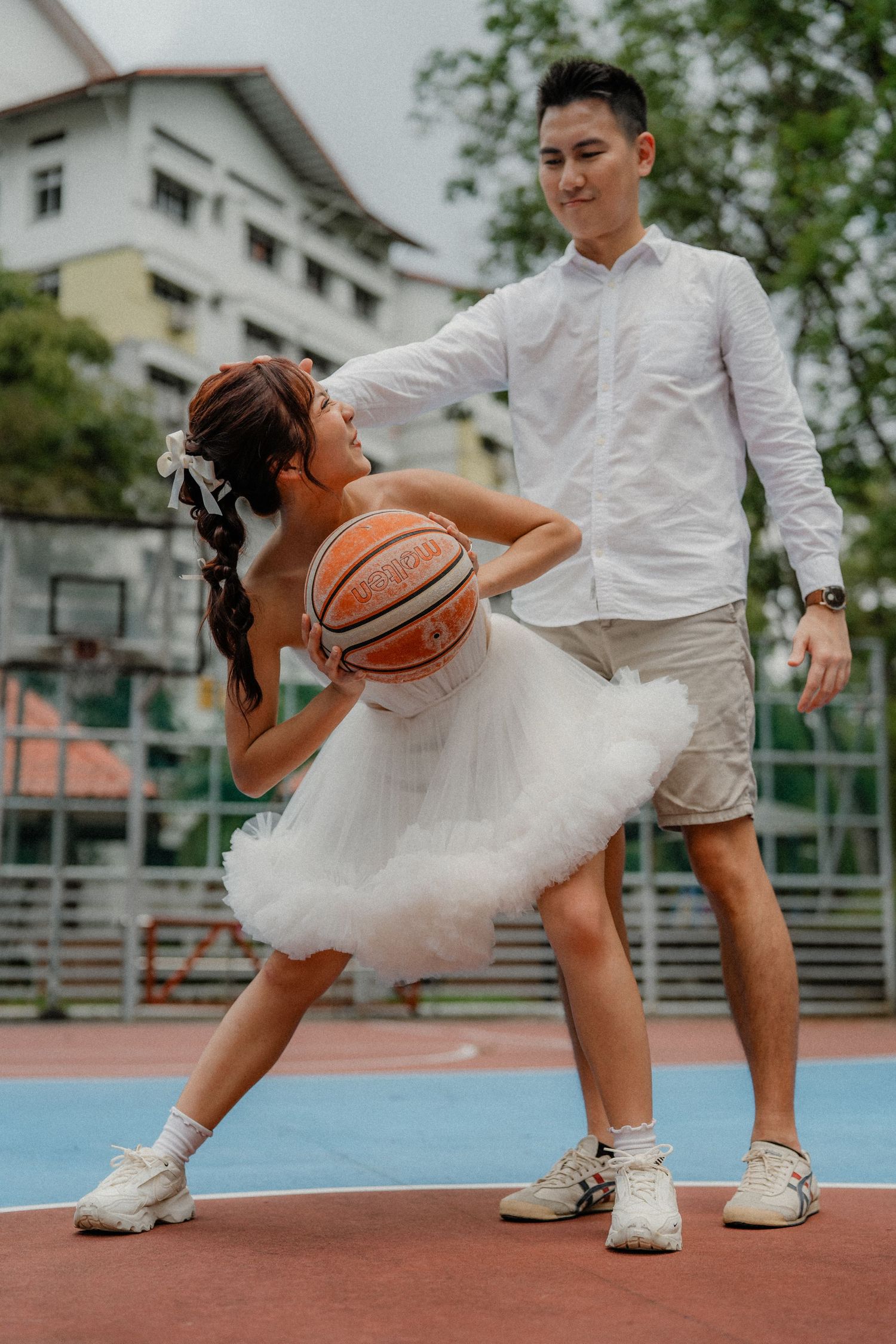 A couple in wedding attire poses playfully on a basketball court with the bride holding a basketball.