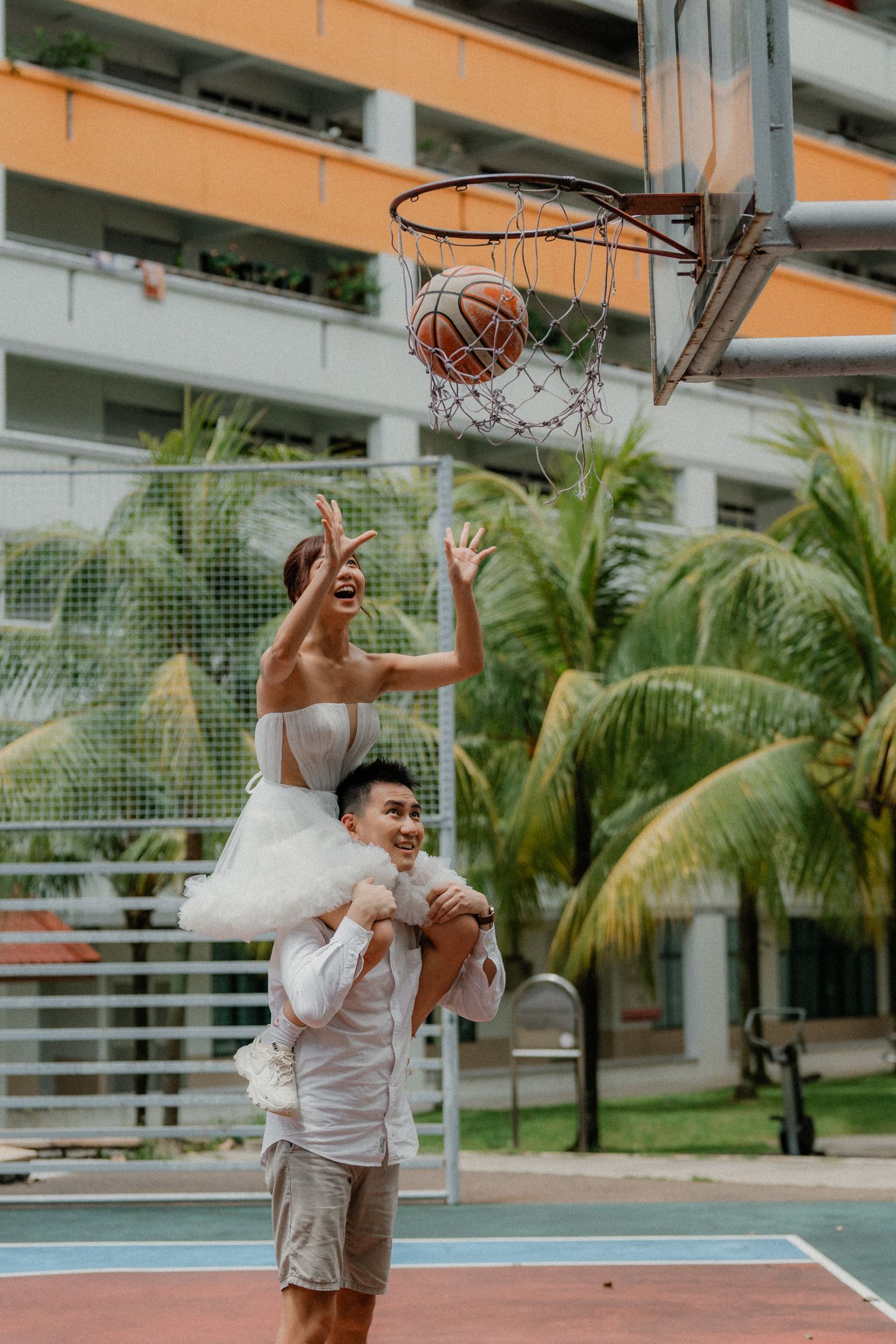 A couple in formal wear playfully shoots hoops on a tropical basketball court with palm trees in the background.