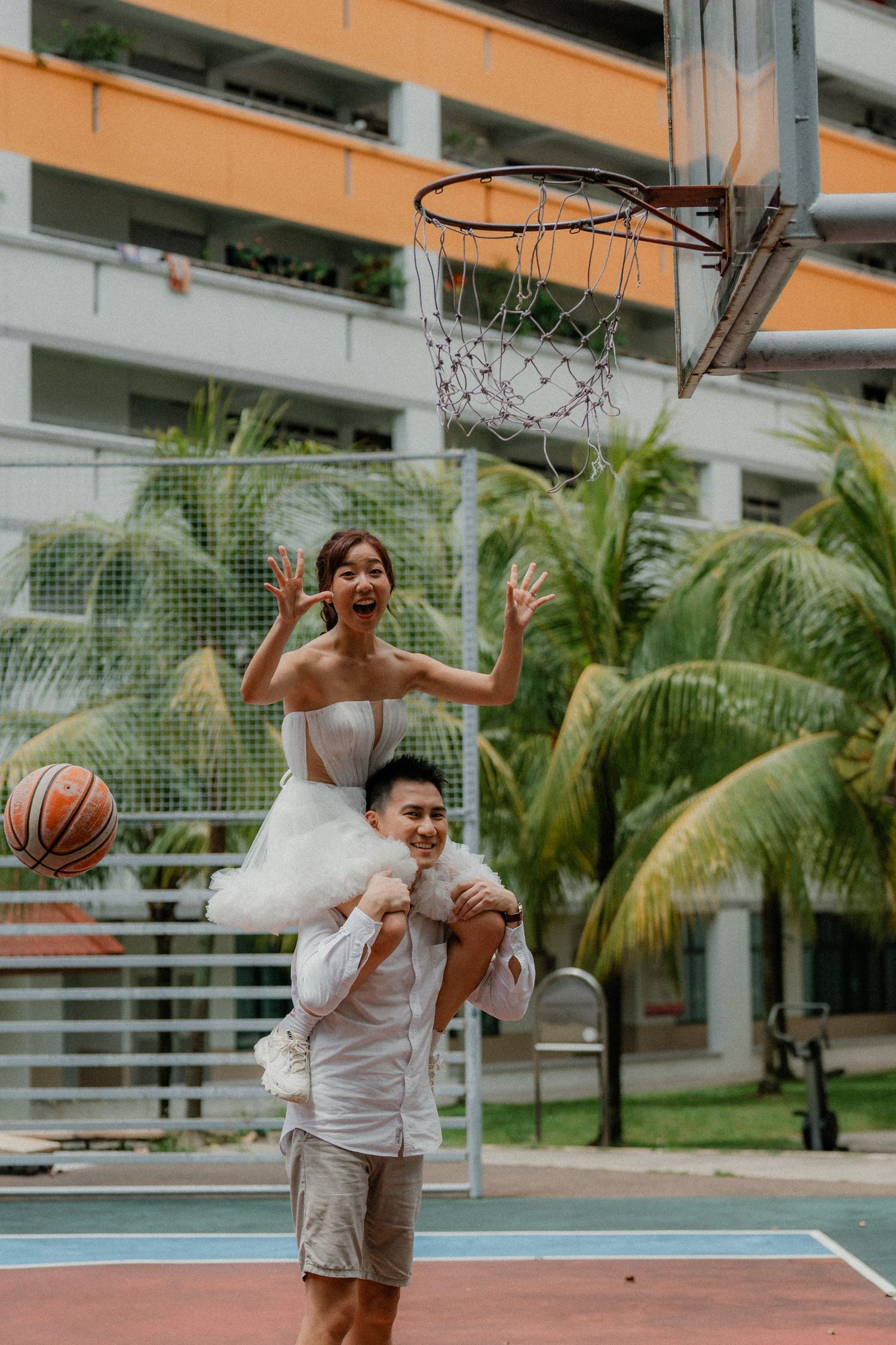 A playful couple poses by a basketball hoop with the bride lifted on shoulders for a fun wedding photo.