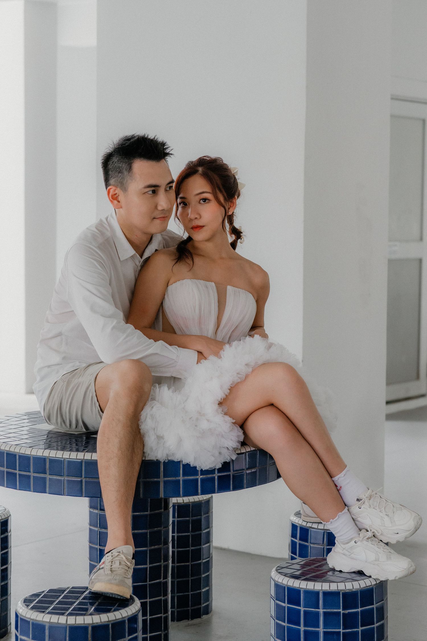 A romantic couple poses intimately on blue mosaic columns while wearing casual white attire and sneakers.
