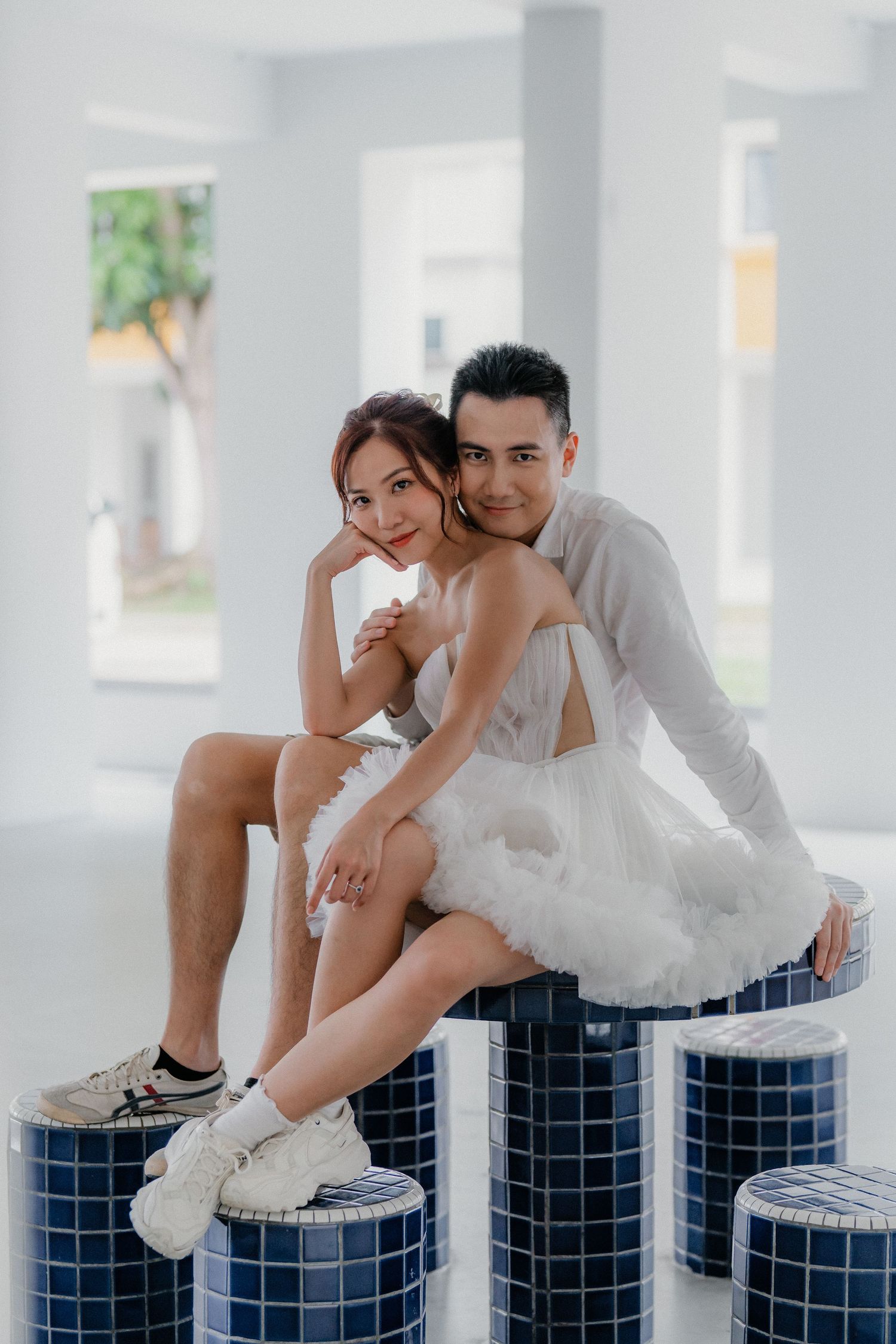 Carefree couple wearing sneakers and white outfits share an intimate moment while sitting on blue mosaic columns.