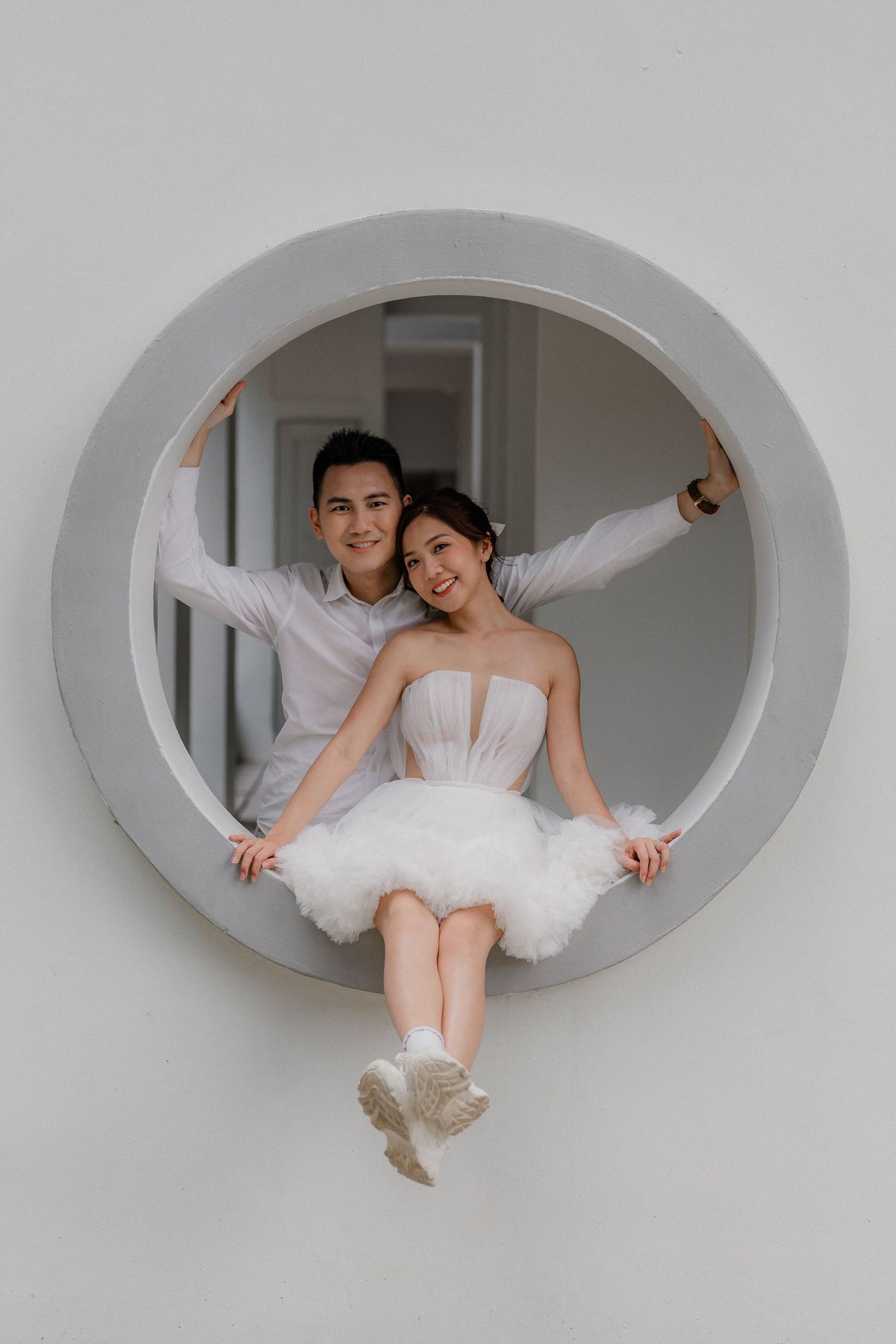 A couple poses together inside a circular window frame against a white wall.