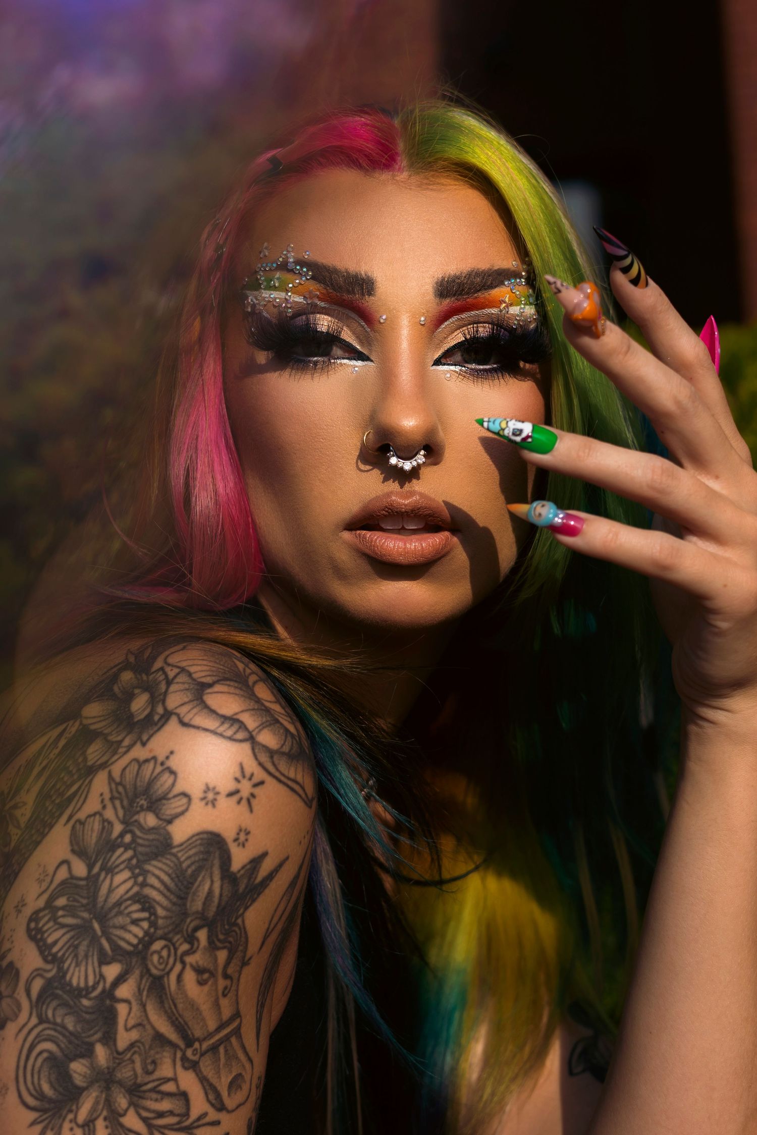 Artistic beauty portrait showing rainbow colored hair, dramatic makeup, shoulder tattoo and glittery eyeshadow against dark backdrop.