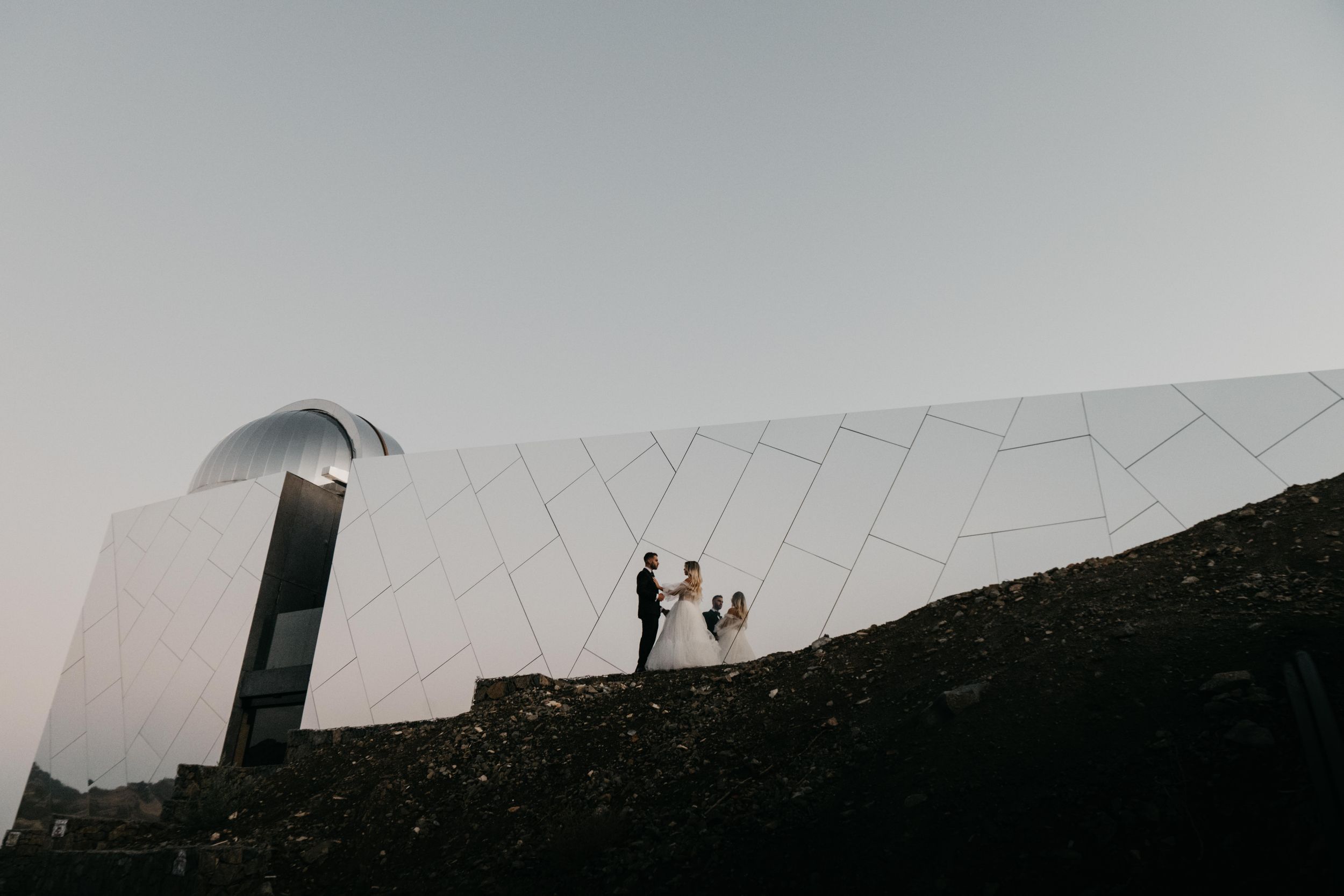 A silhouetted couple stands against a modern white geometric structure on a dark hill during twilight.