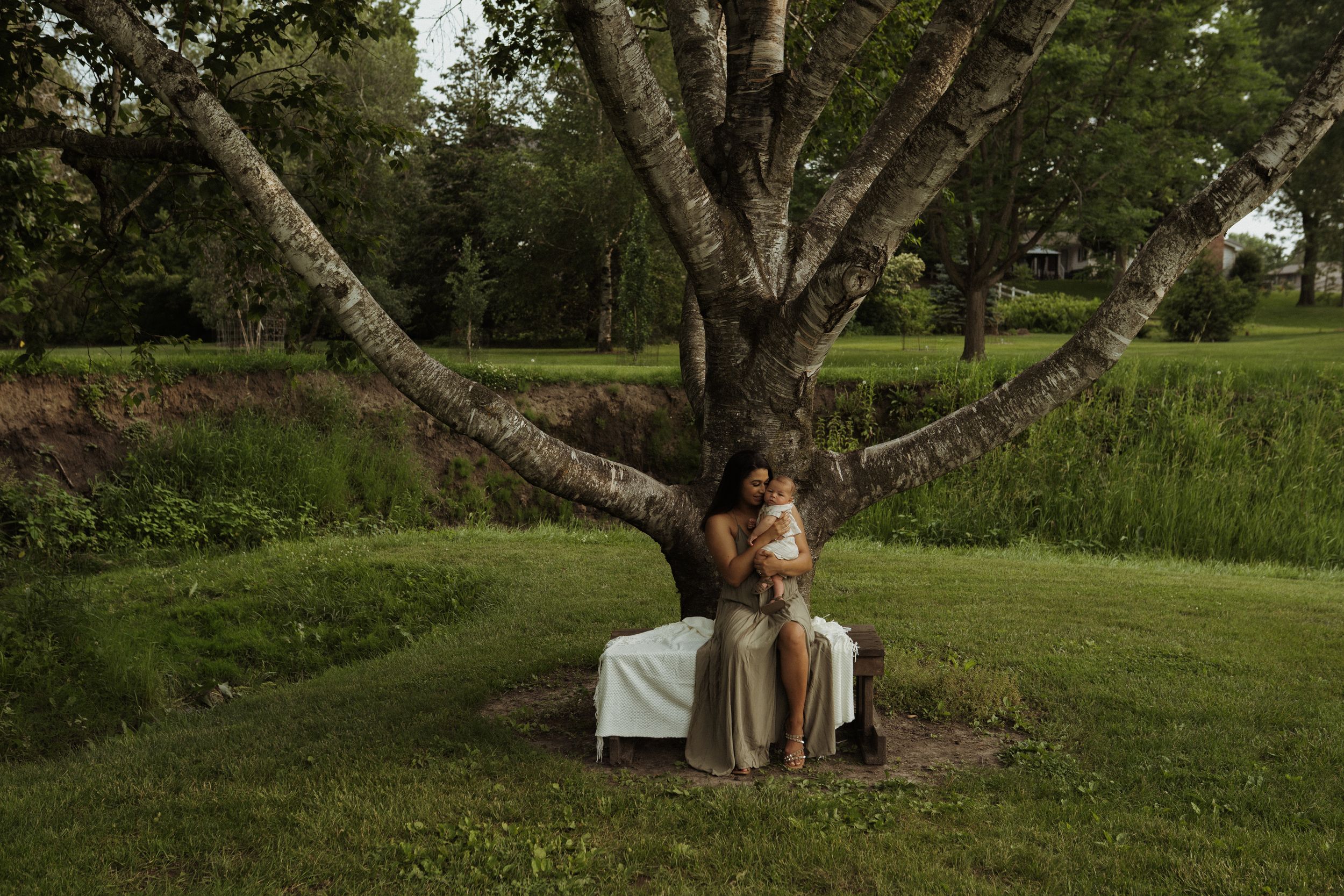 A person in a flowing dress stands beneath a large branching tree in a lush green setting.